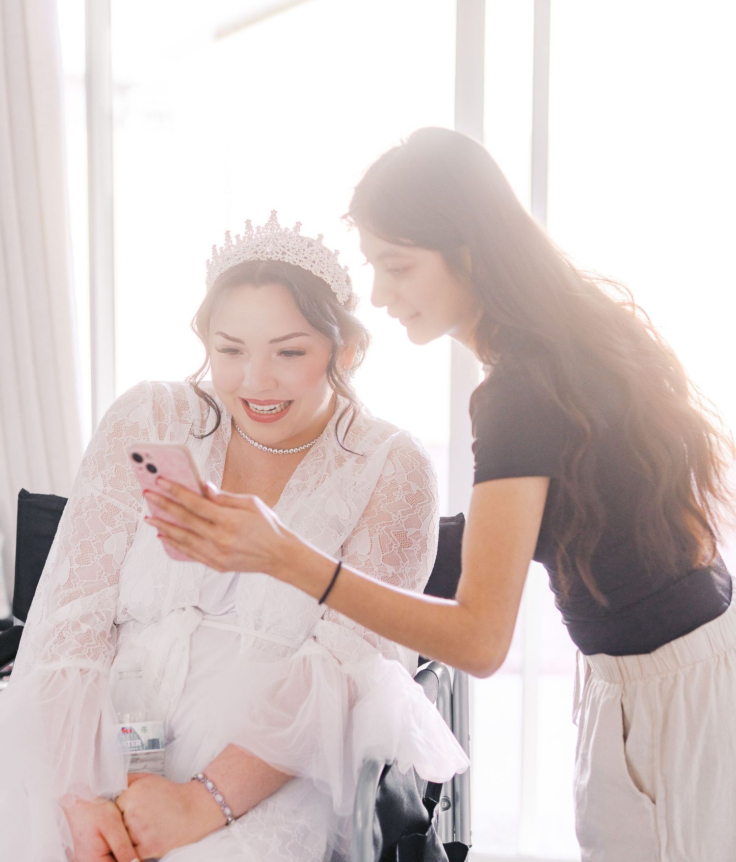 Two people looking at a smartphone together in a brightly lit room with white curtains in the background.