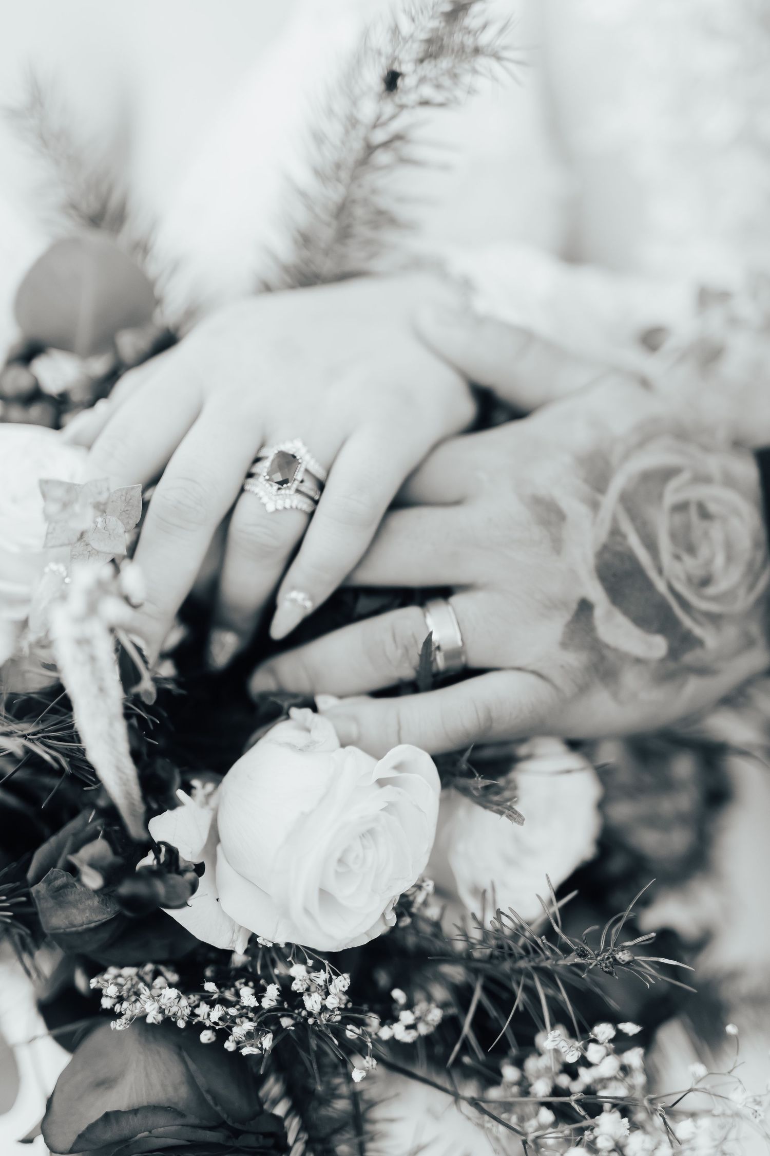 Black and white photograph showing wedding rings and delicate roses in a romantic bridal bouquet arrangement.