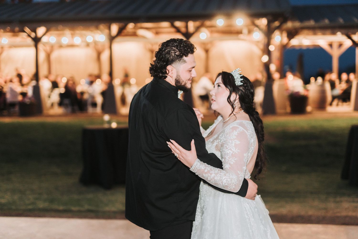 A couple shares a romantic dance moment at an outdoor evening reception under warm string lights and a covered pavilion.
