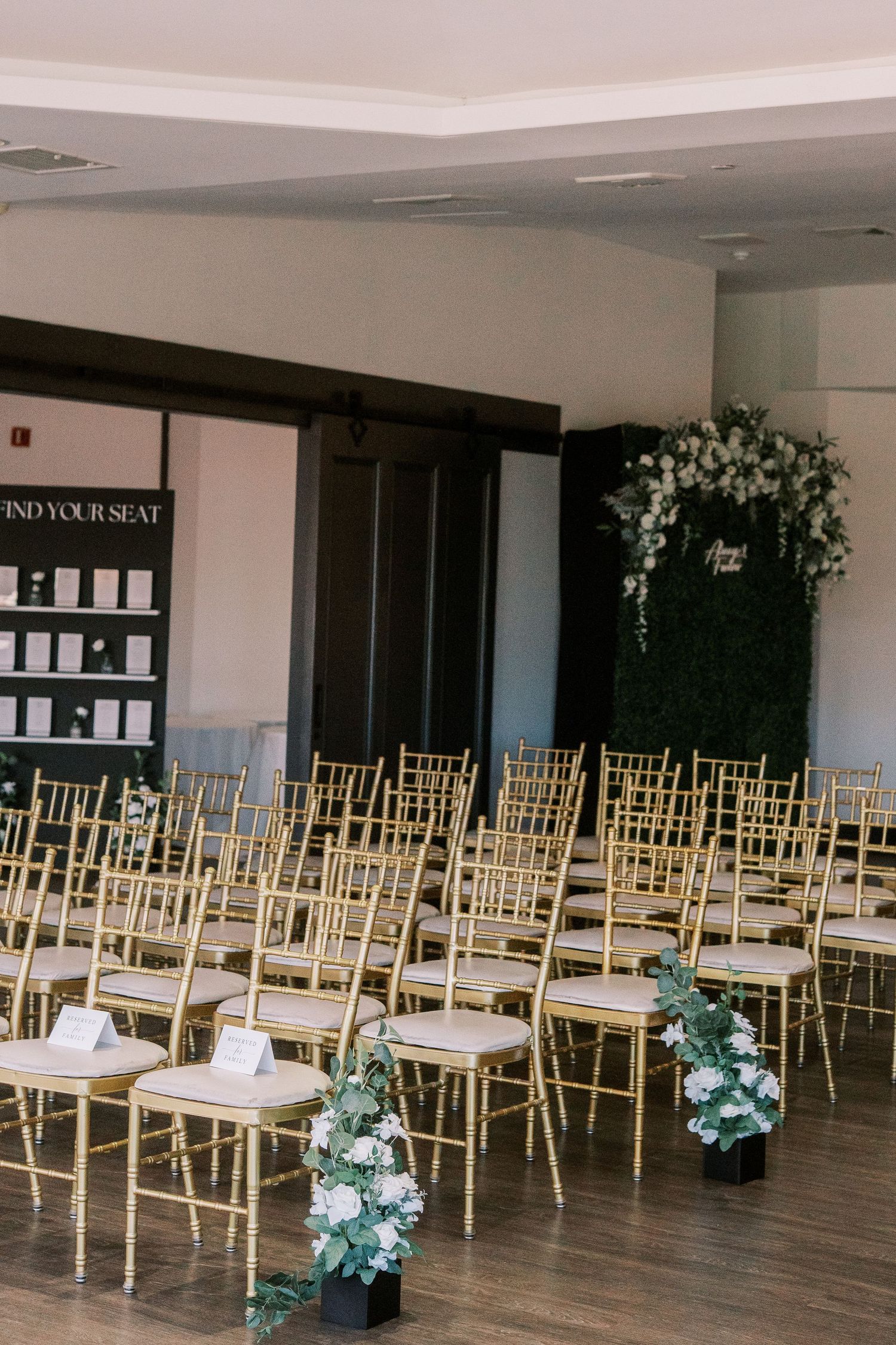 Wedding ceremony setup with rows of gold chiavari chairs and green floral arrangements in an elegant indoor venue.