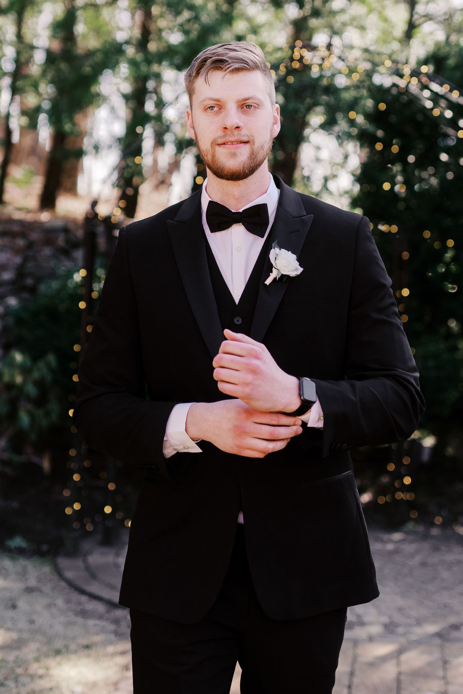 A formal portrait of a groom in a classic black tuxedo and bowtie with twinkle lights behind him.