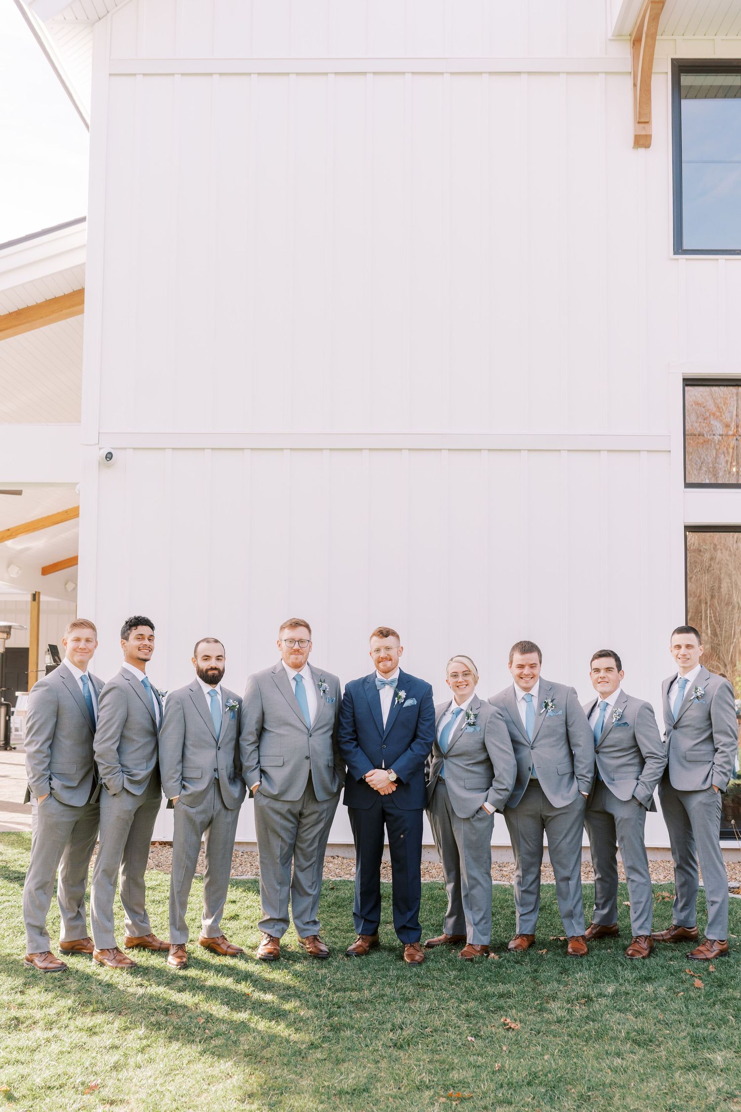Wedding party members in matching gray suits and brown shoes pose together on grass outside a white building.