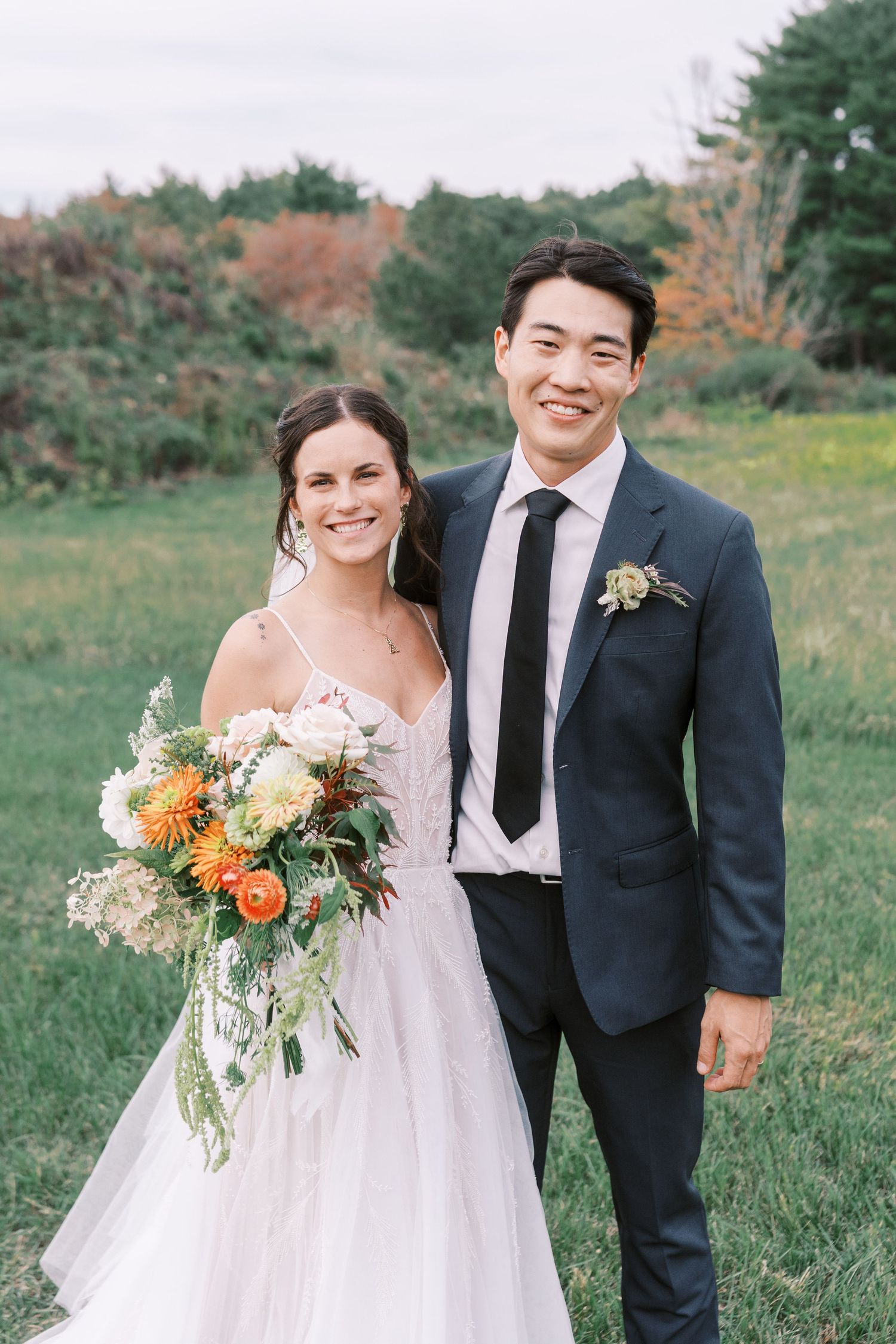 A wedding couple poses together in a grassy field with the bride holding a vibrant floral bouquet.