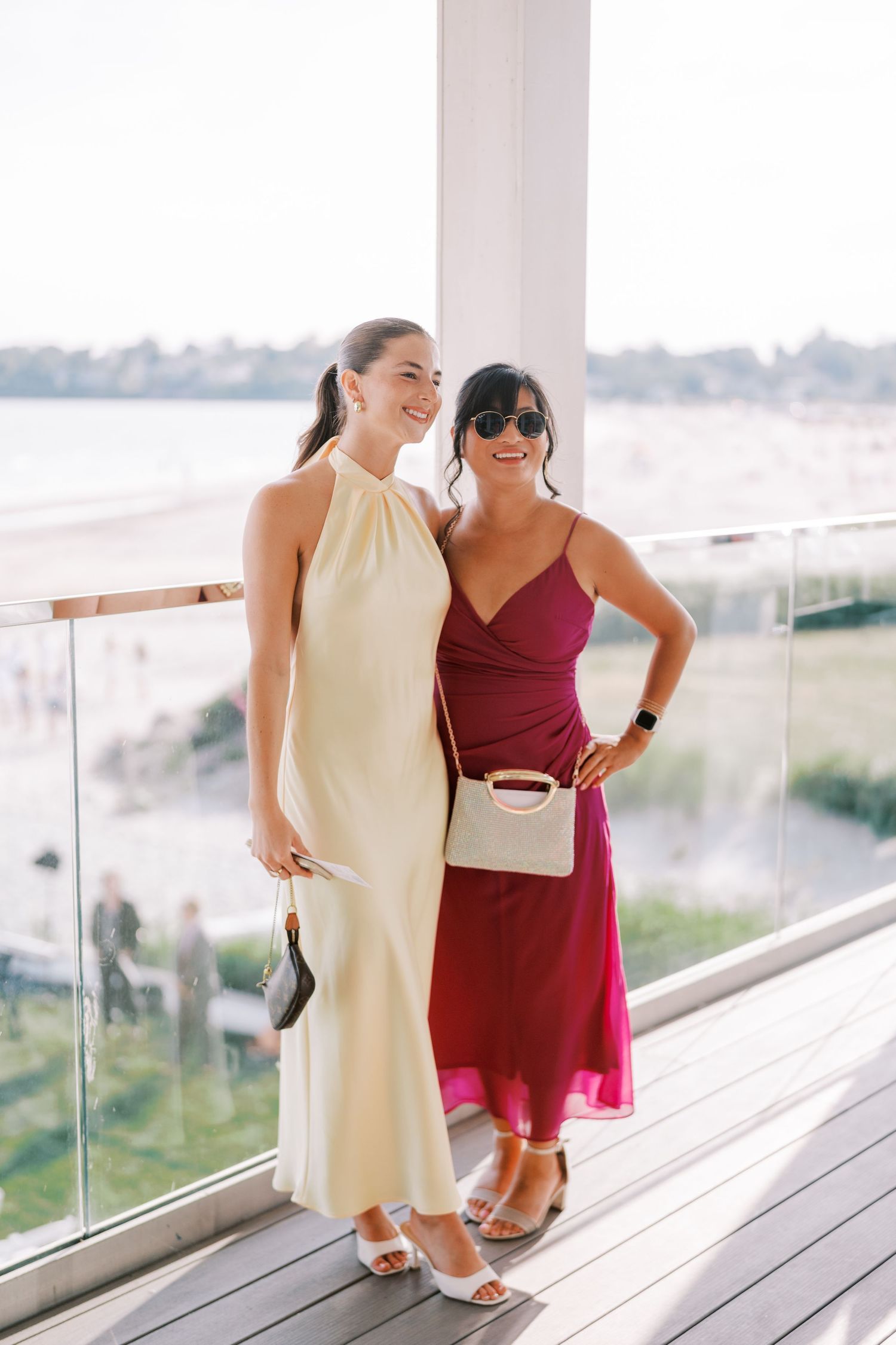 Two women in contrasting dresses laugh together on a coastal deck with glass railings and ocean views.