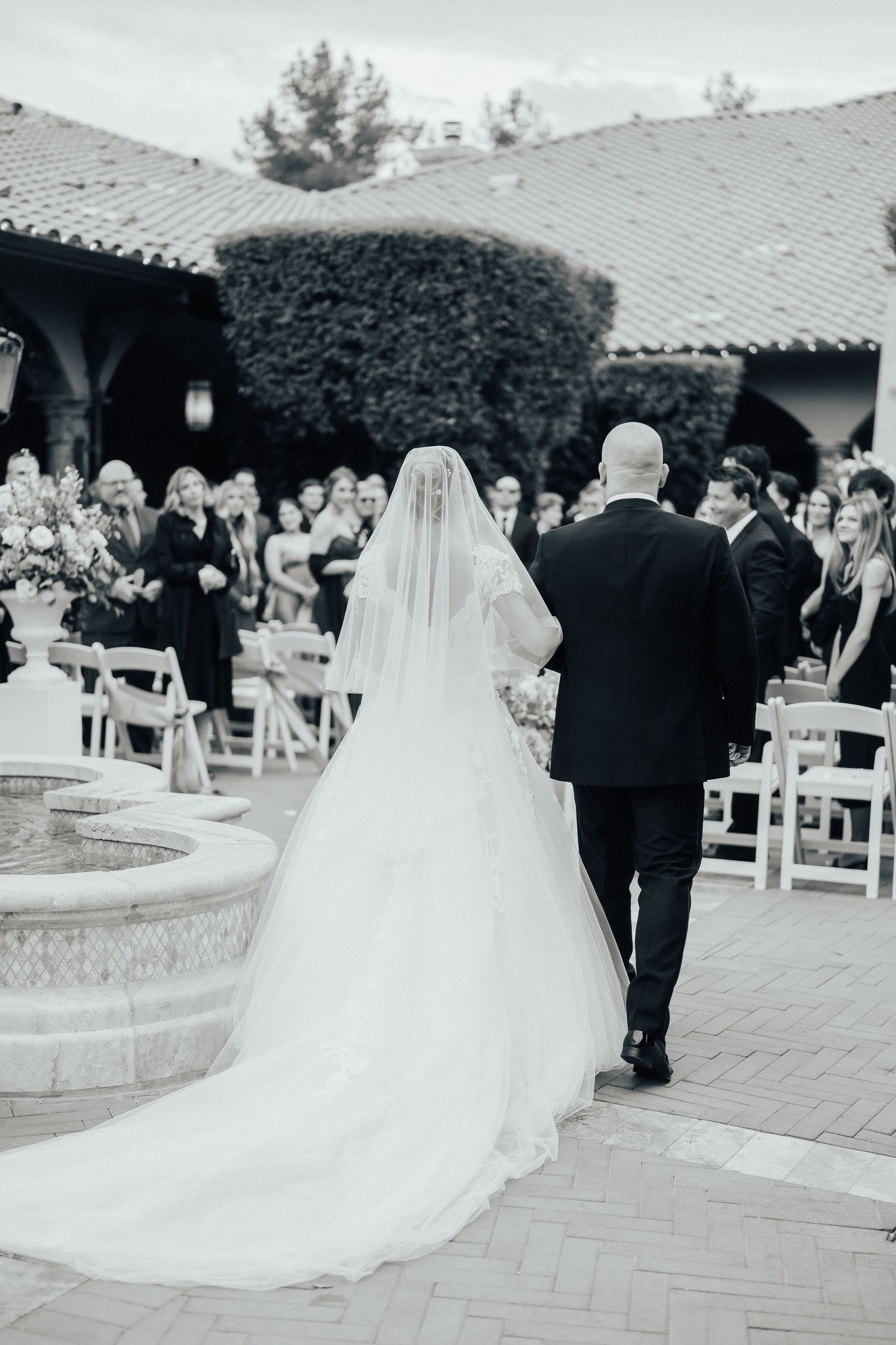 A wedding ceremony at an outdoor venue with a long flowing white veil and dress trailing behind on brick pavers.