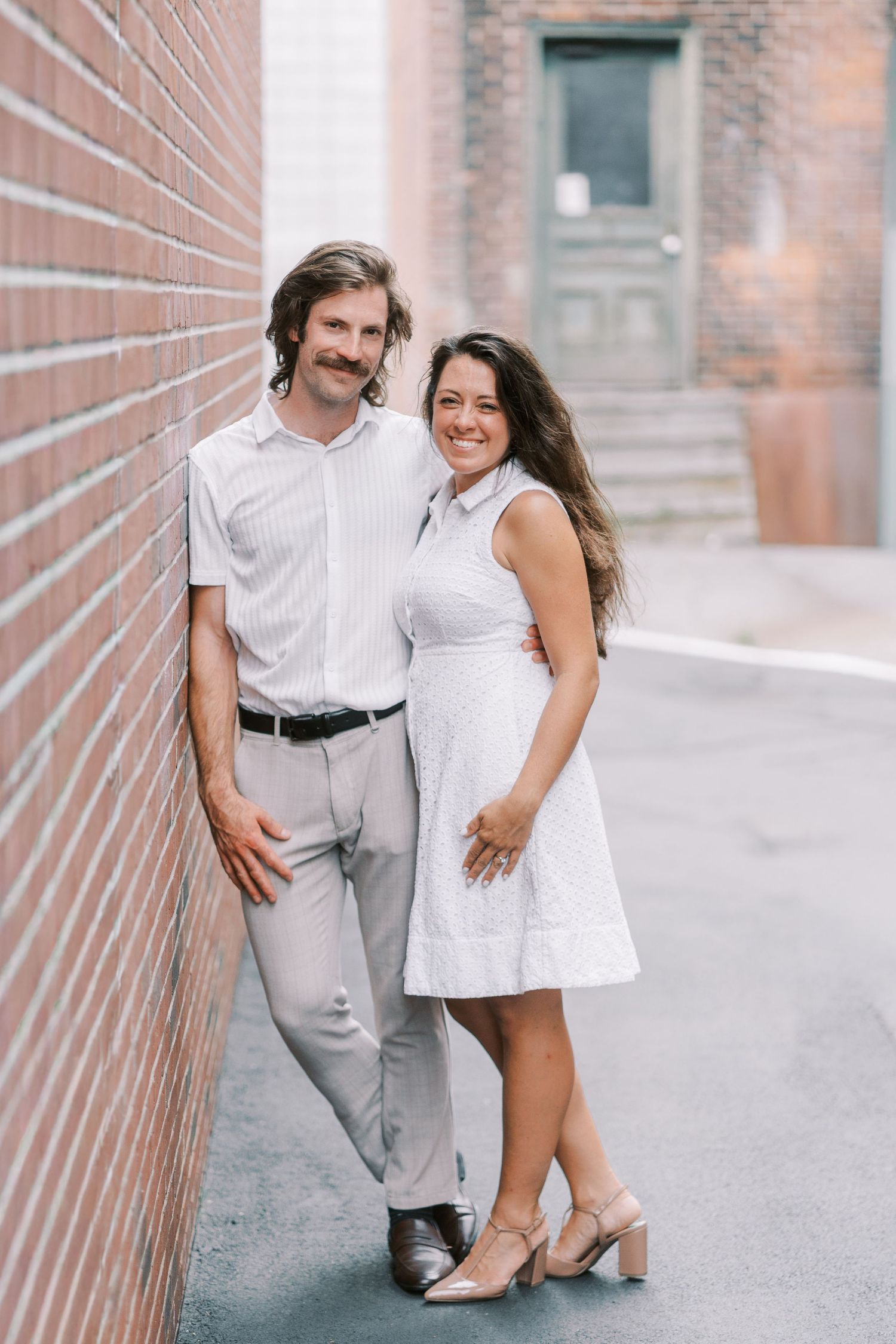 A stylish couple in white attire poses against a brick wall backdrop in an urban setting.