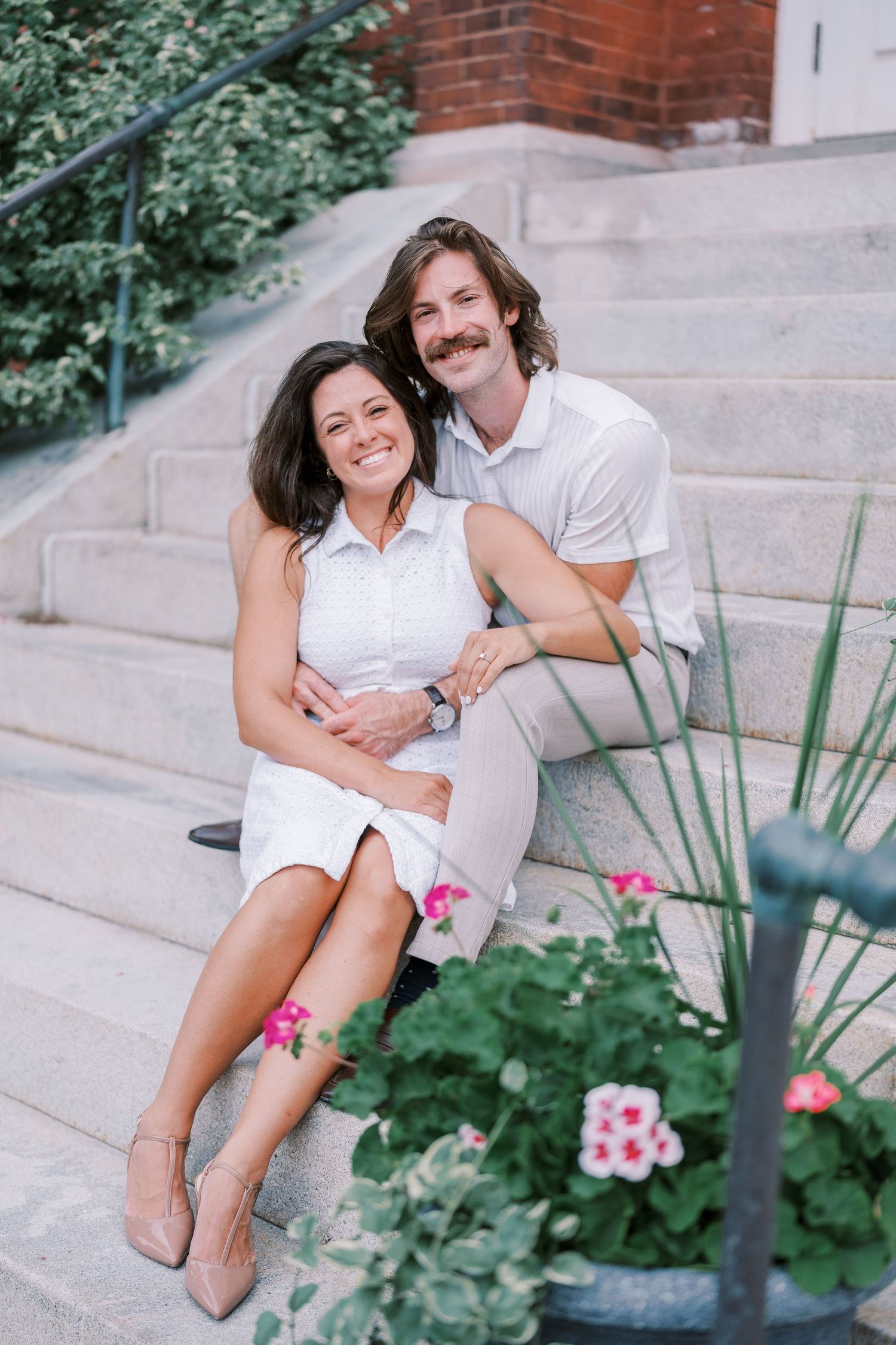 A couple in coordinating white outfits sits together on concrete steps with pink flowers blooming nearby.
