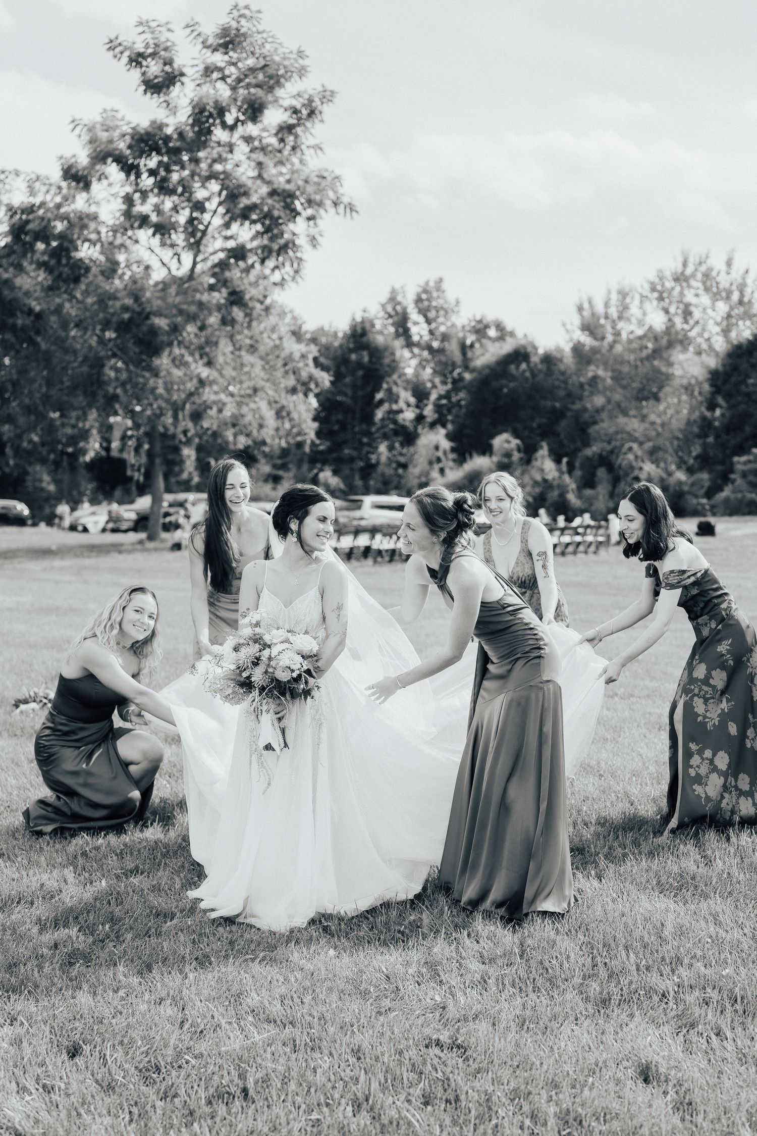 Black and white photo of bridesmaids helping arrange wedding dress outdoors.