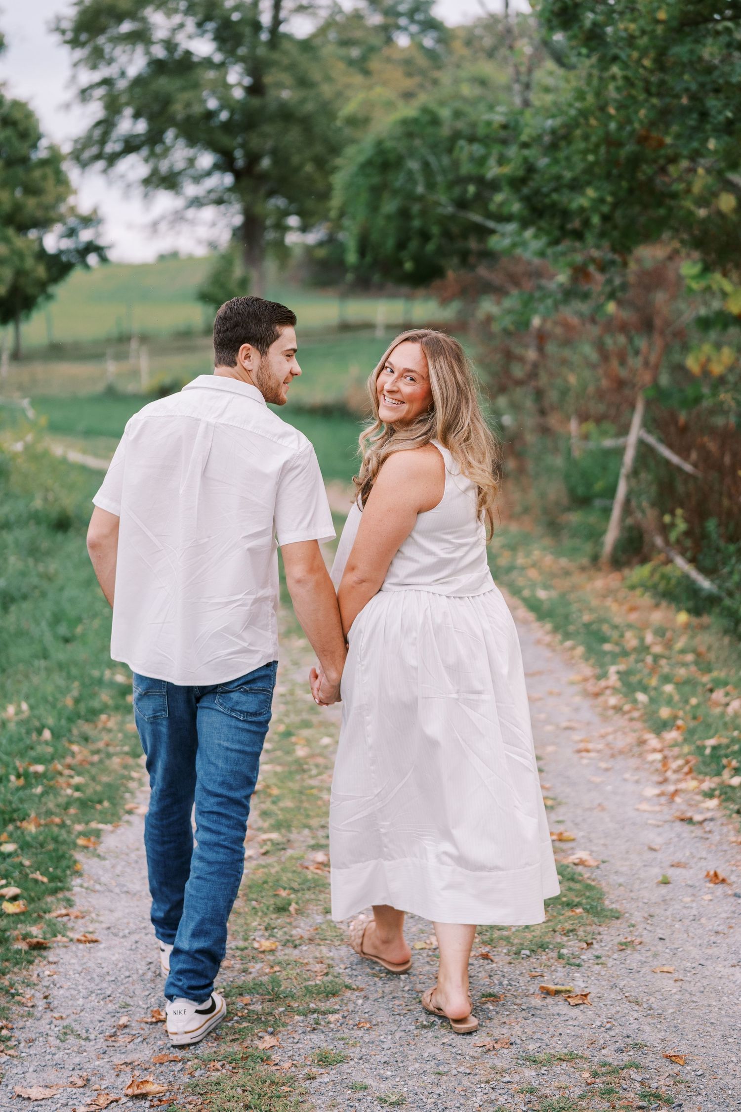 Couple in coordinating white outfits walking hand in hand down rural path.