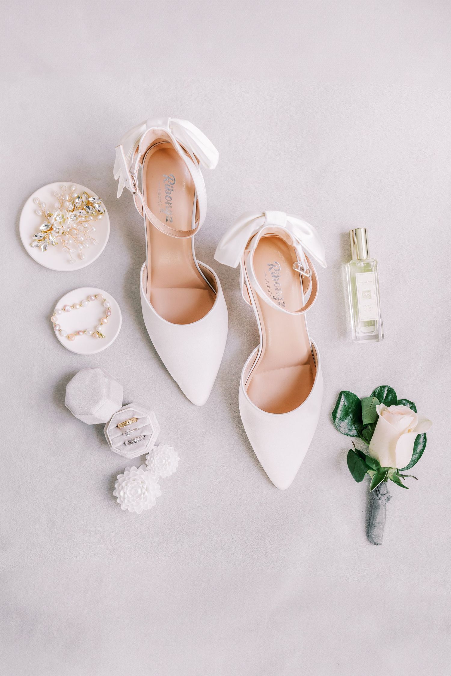 Elegant white bridal shoes displayed with perfume, jewelry, and a delicate green boutonniere.