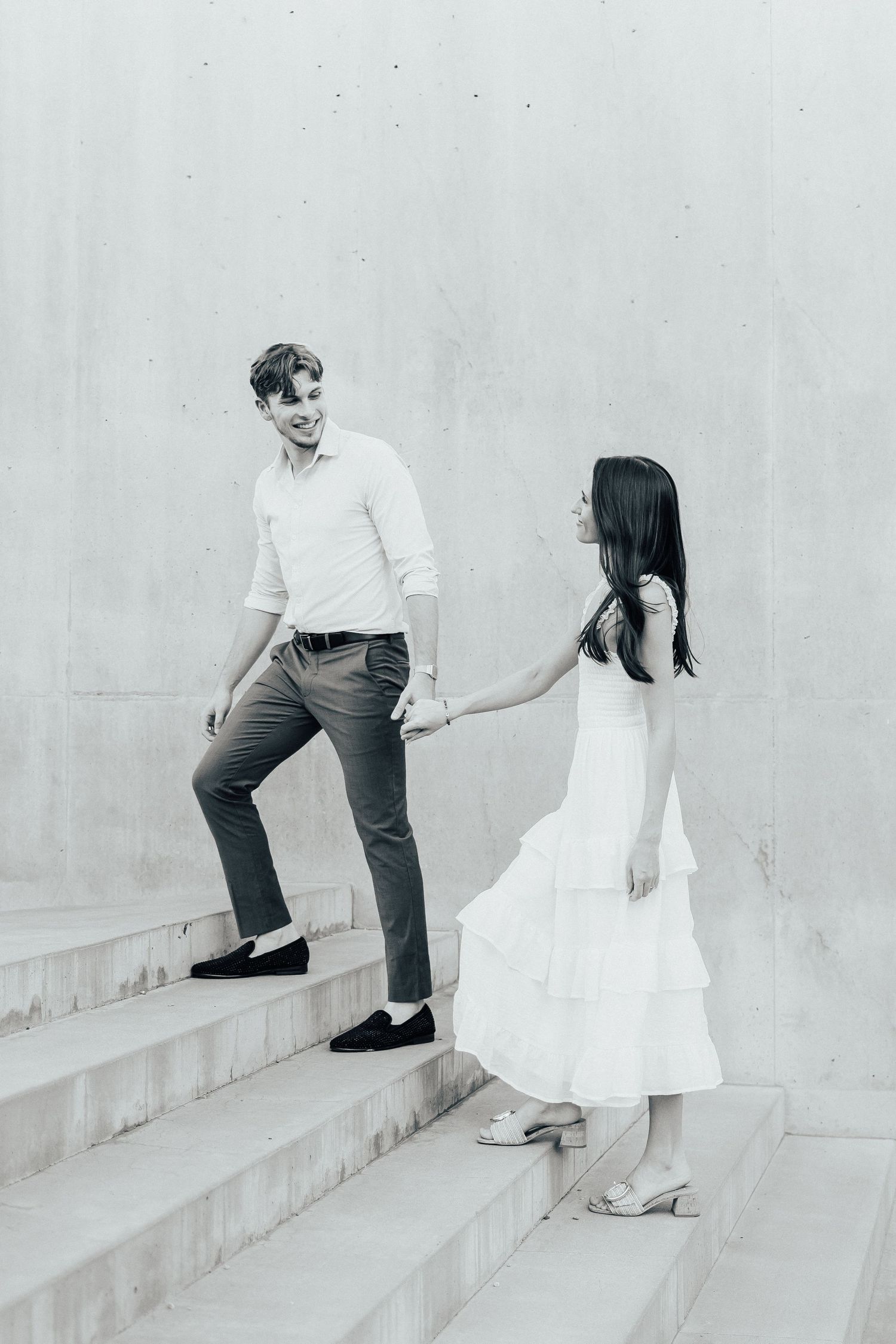 A couple posing on concrete steps in black and white, with the woman in white dress and man in dress pants.