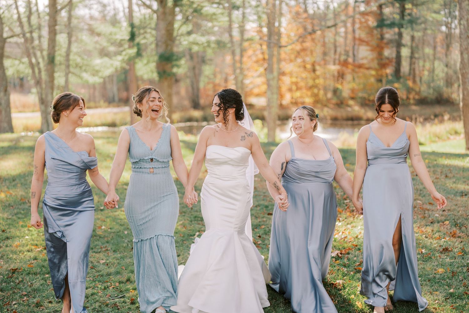 Bridesmaids in dusty blue dresses walk together through a sunlit autumn forest during an outdoor wedding celebration.