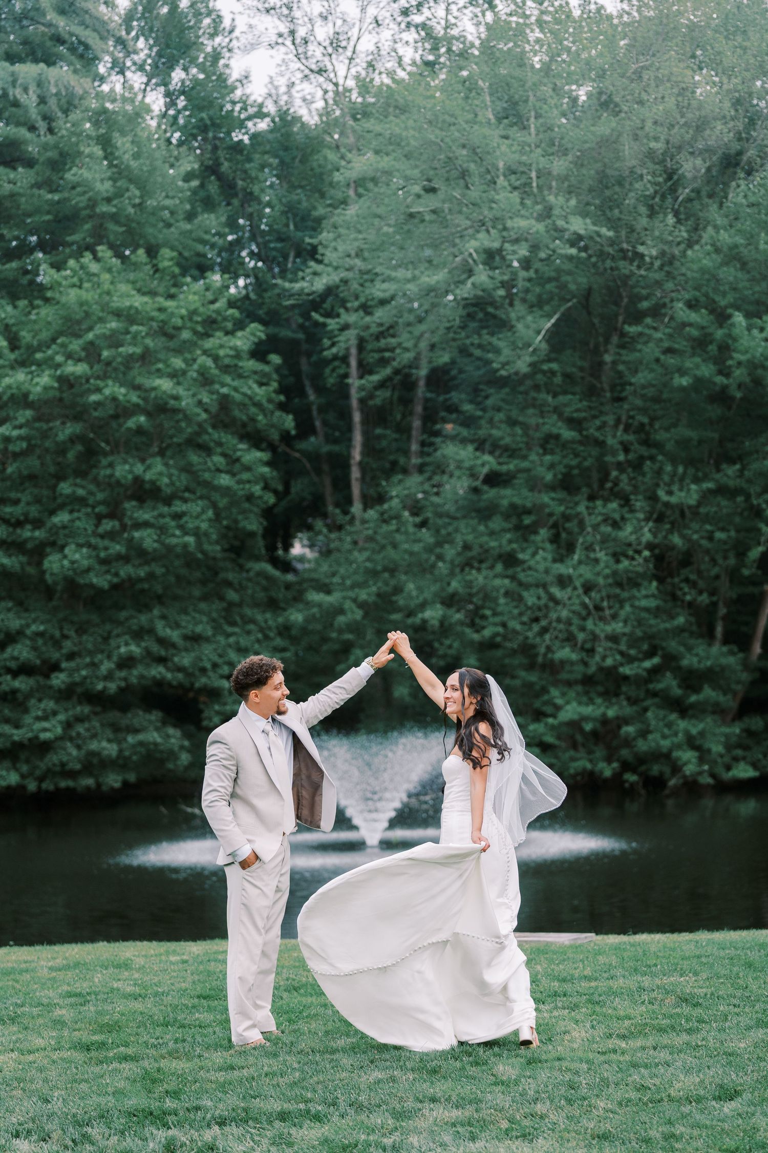 A bride and groom dance together on a grassy lawn beside a serene lake surrounded by lush forest.