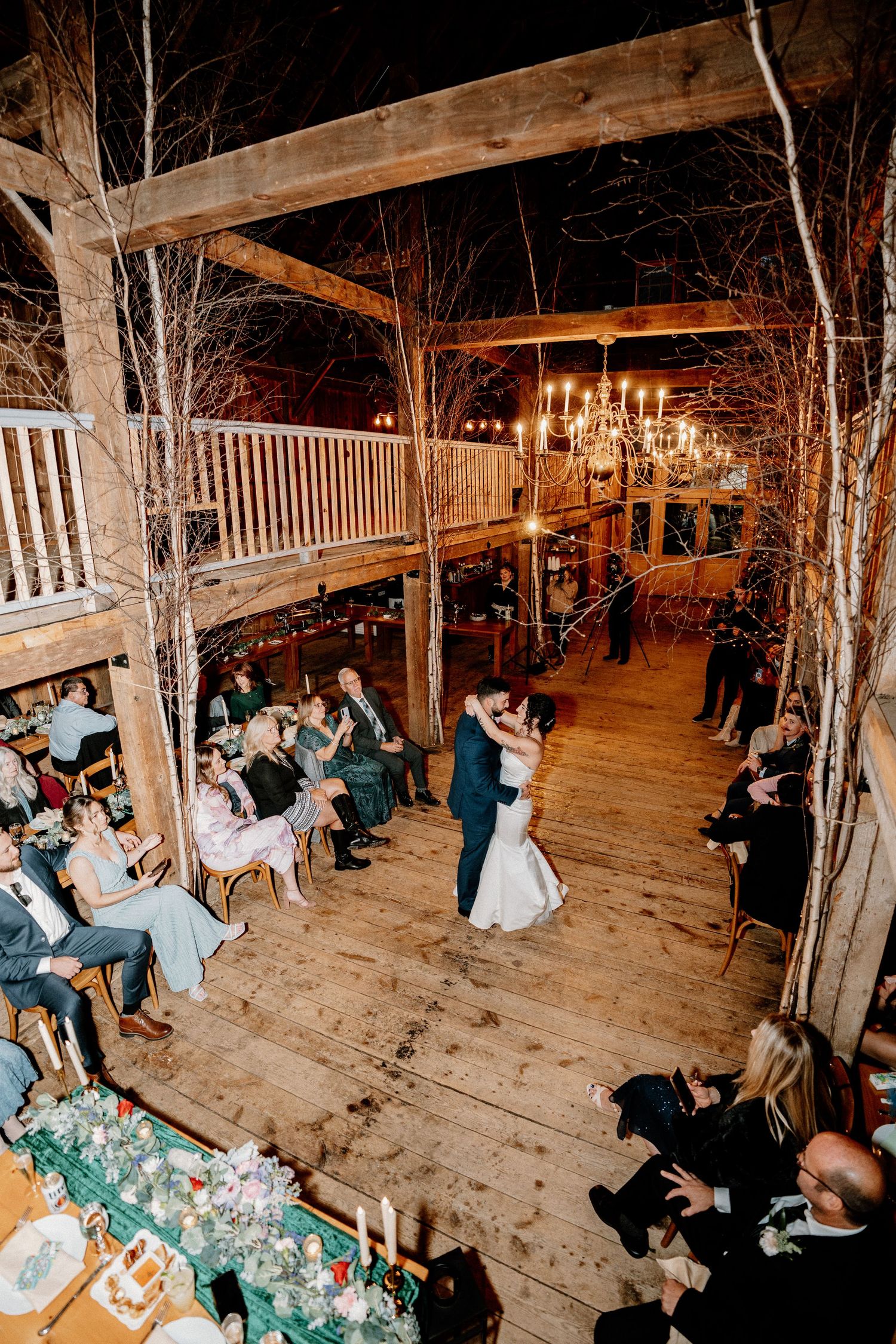 Rustic barn wedding reception venue with wooden beams, chandeliers and guests seated at tables during evening celebration.
