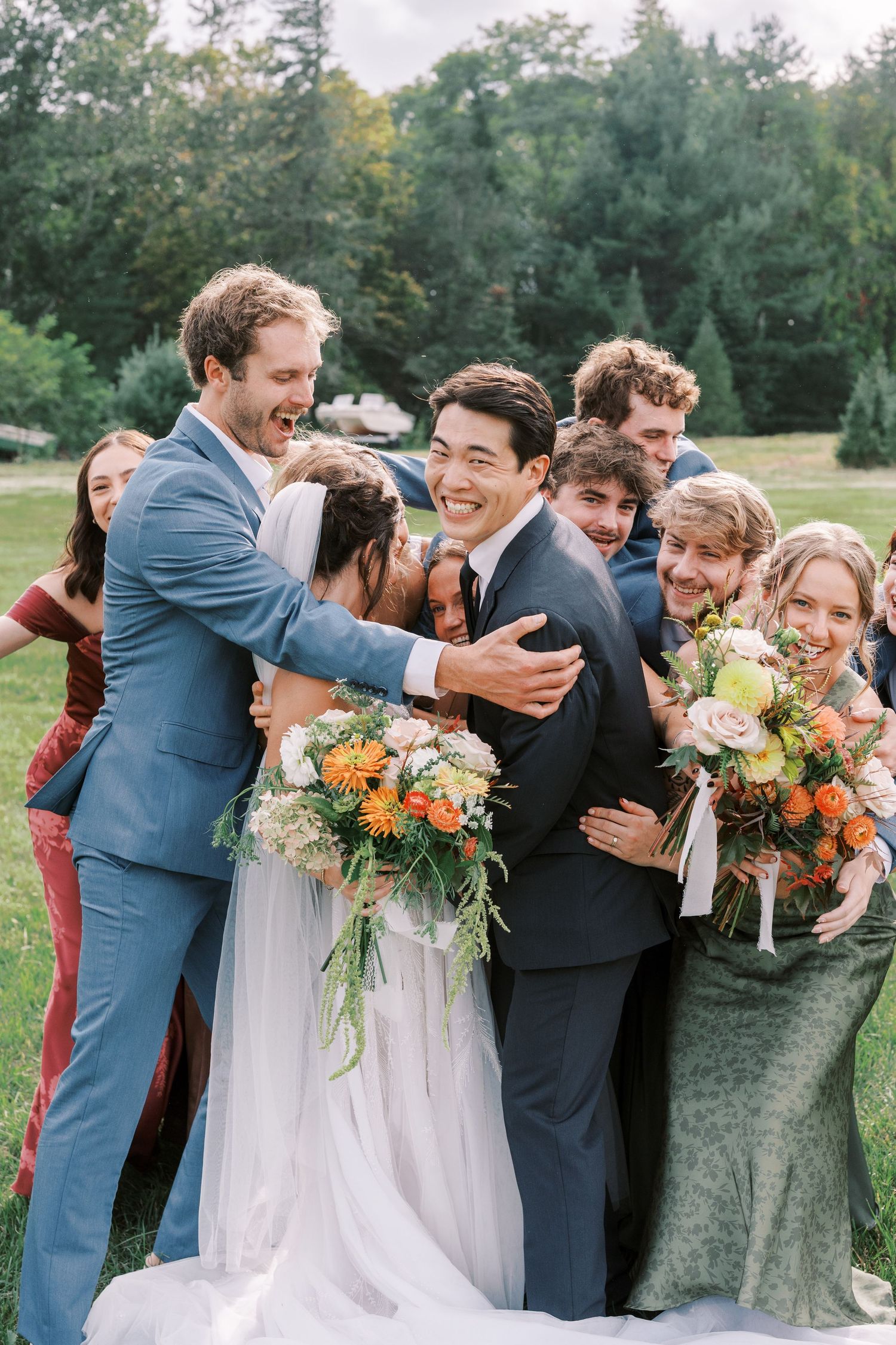 Wedding party laughing and embracing outdoors with colorful bouquets and blue suits.