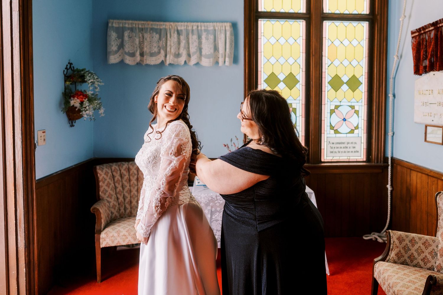 A bride and bridesmaids get ready in a church hallway with ornate stained glass windows and red carpet.