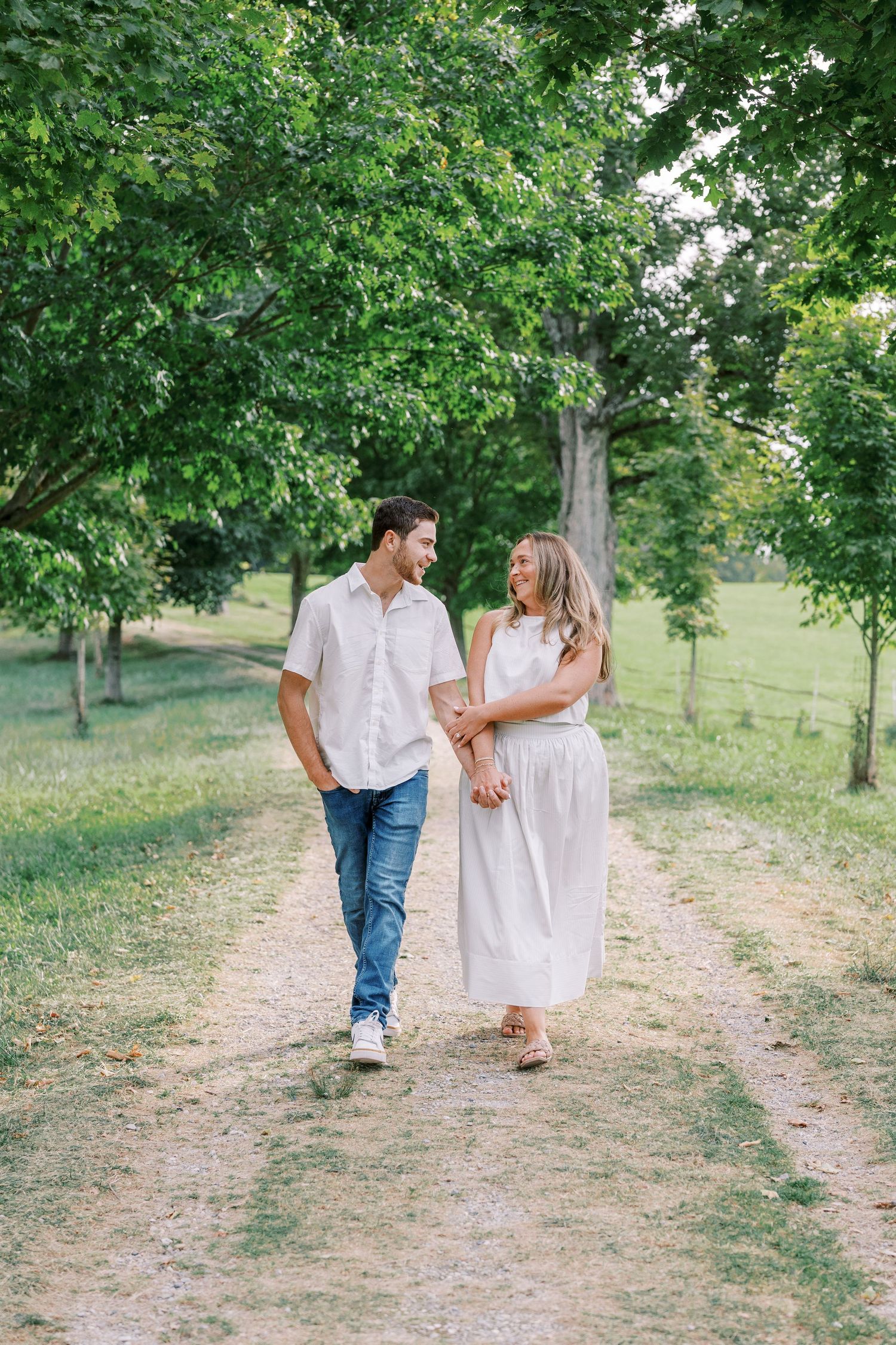 A couple walks hand in hand down a scenic path surrounded by green trees.