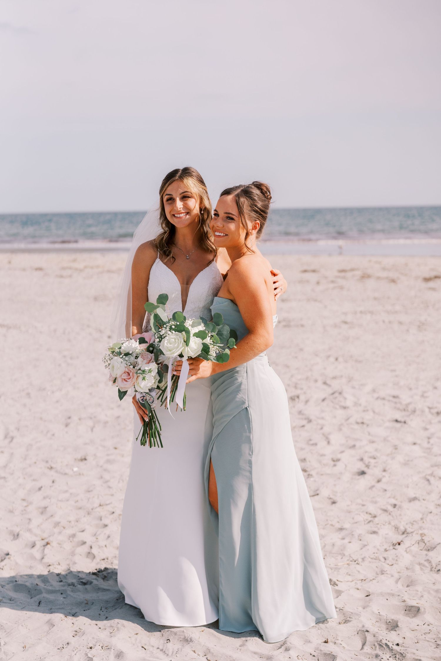 Two people in formal attire share an emotional moment on a sunny beach with white sand and blue water in the background.