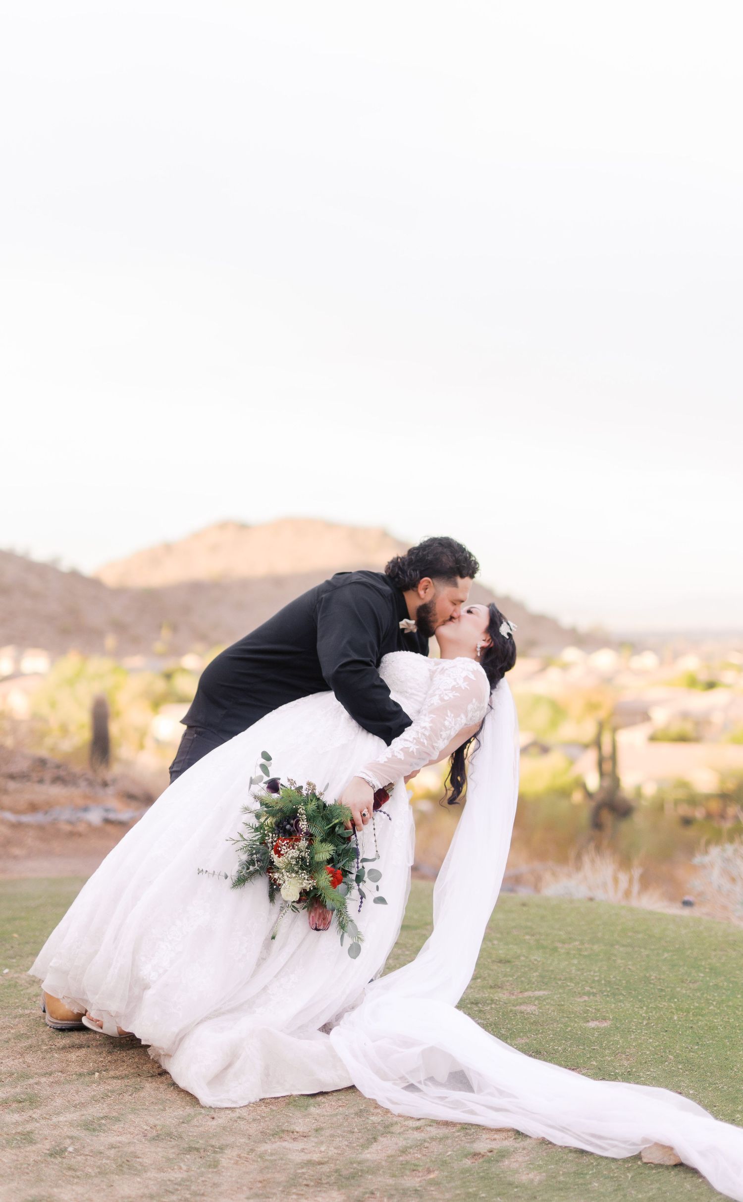 A romantic wedding moment captured in desert landscape as a couple shares a kiss with mountains and cacti in the background.