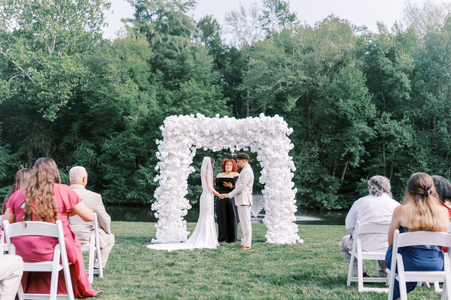 Outdoor wedding ceremony under a beautiful white floral arch with guests seated on white chairs.