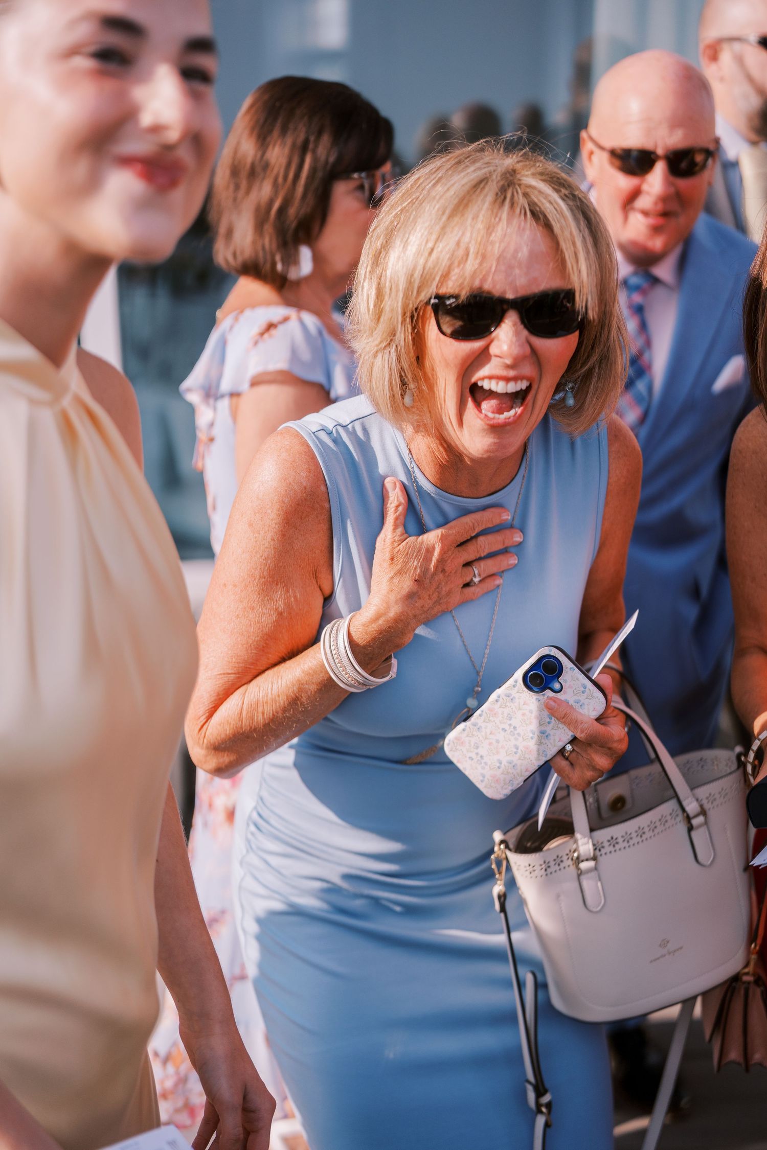 A group of people laughing and socializing at an outdoor event, with someone wearing a light blue dress and white purse.