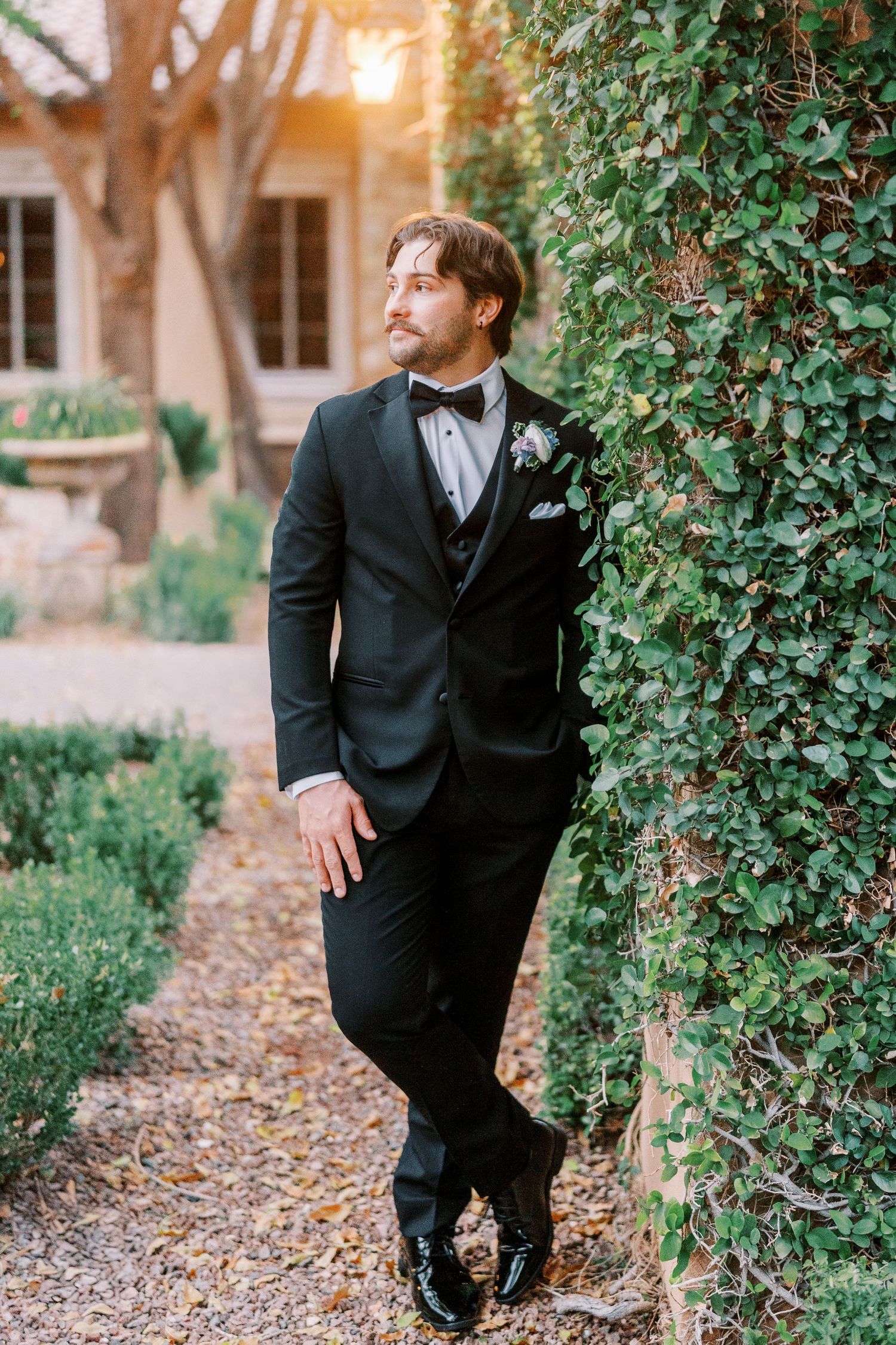 A person in a formal black tuxedo poses against a backdrop of green foliage and stone architecture at sunset.