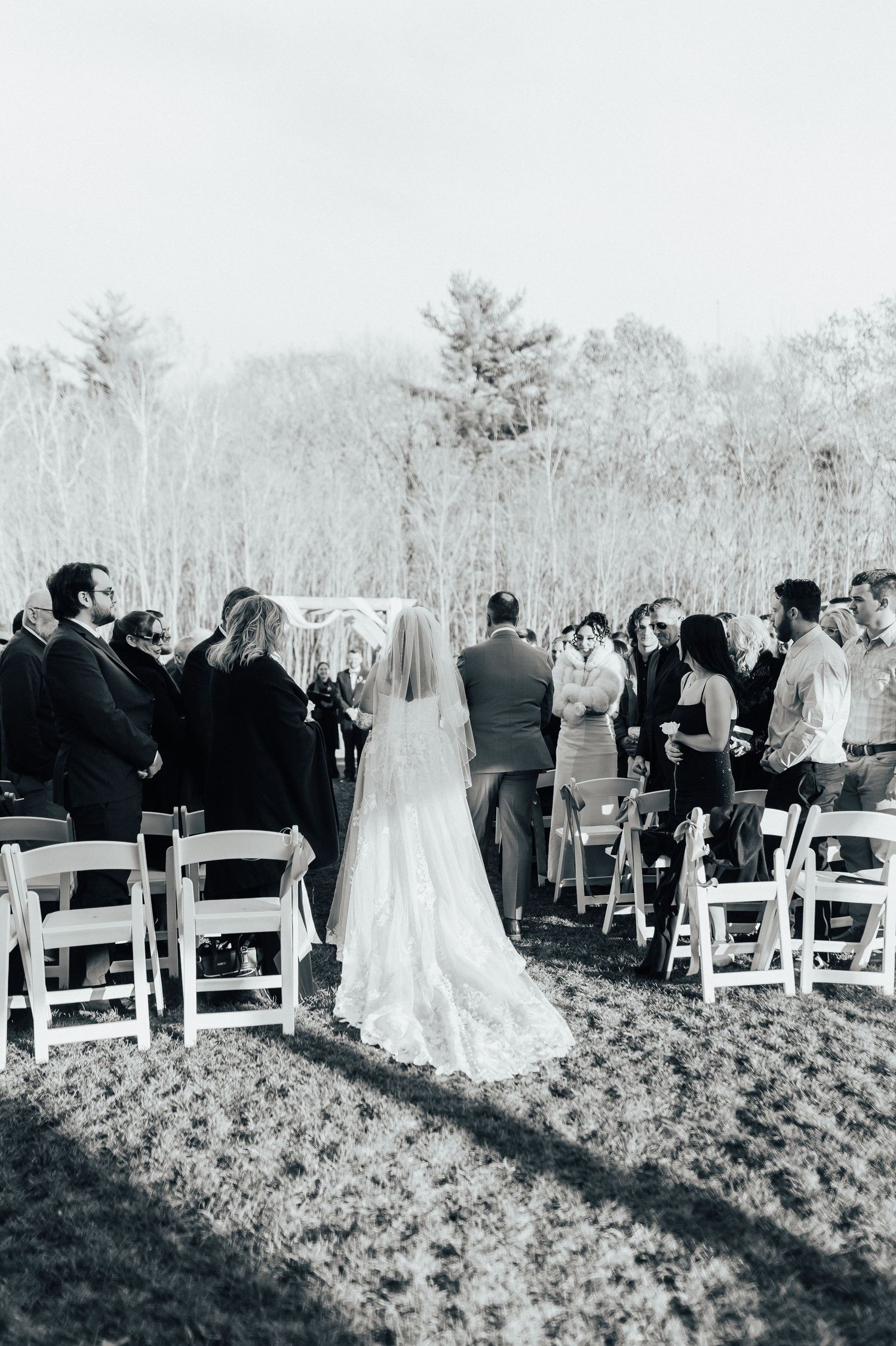 Wedding guests stand and watch outdoor ceremony in black and white as shadows stretch across field.