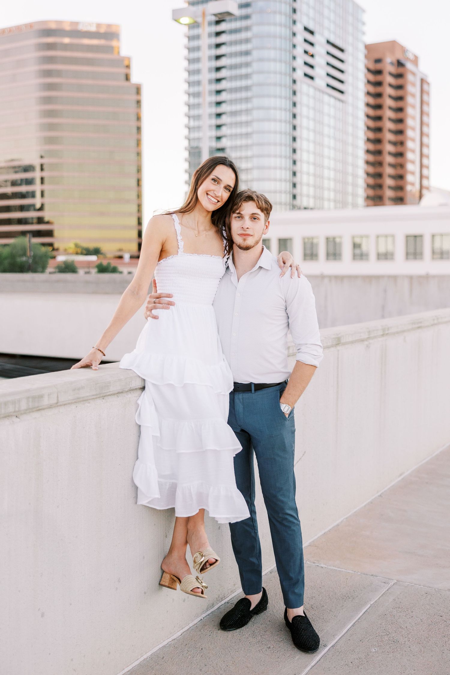 A couple poses playfully on a rooftop terrace with city high-rises in the background.