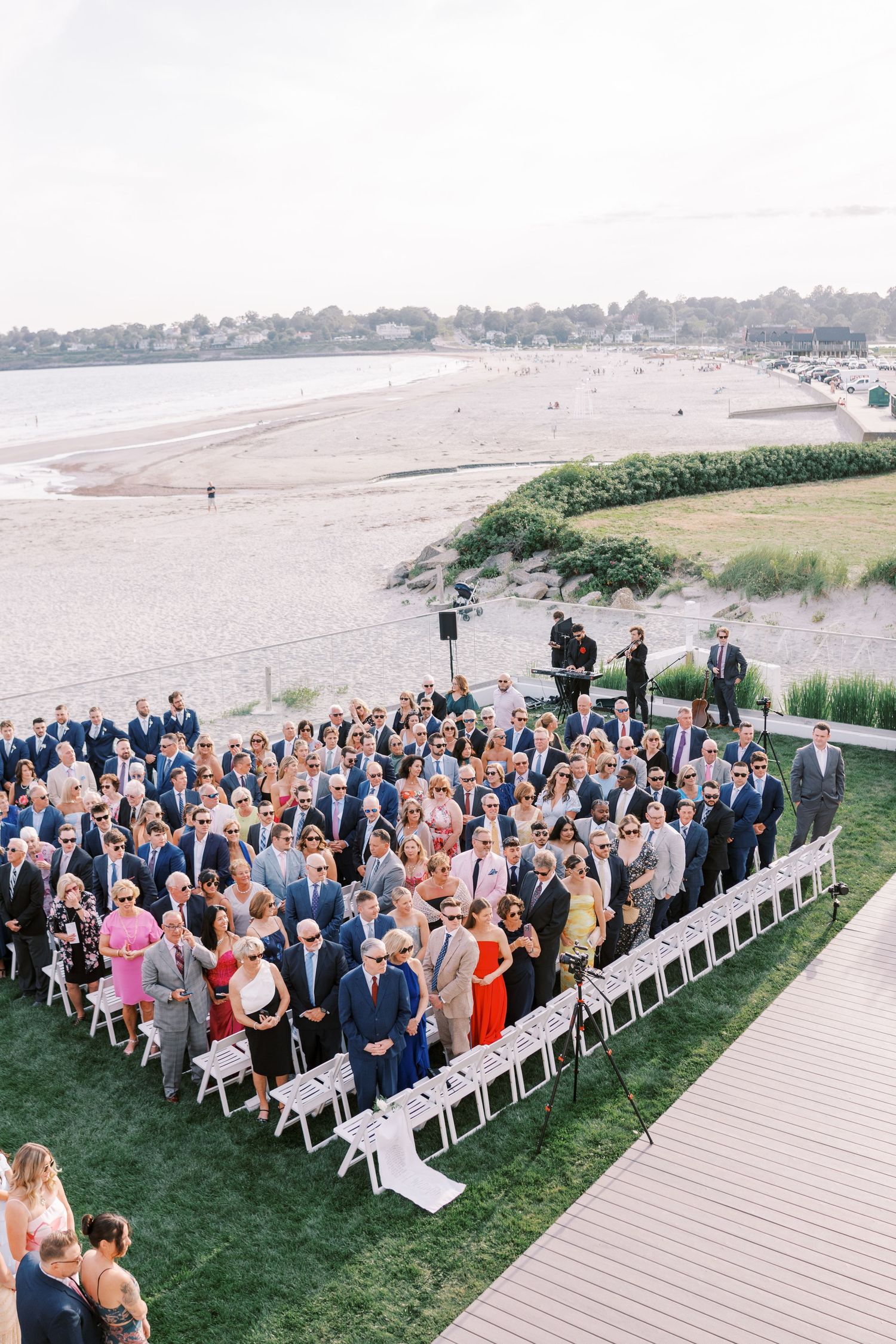 Wedding guests seated in white chairs on a lush green lawn overlooking a scenic waterfront during an outdoor ceremony.