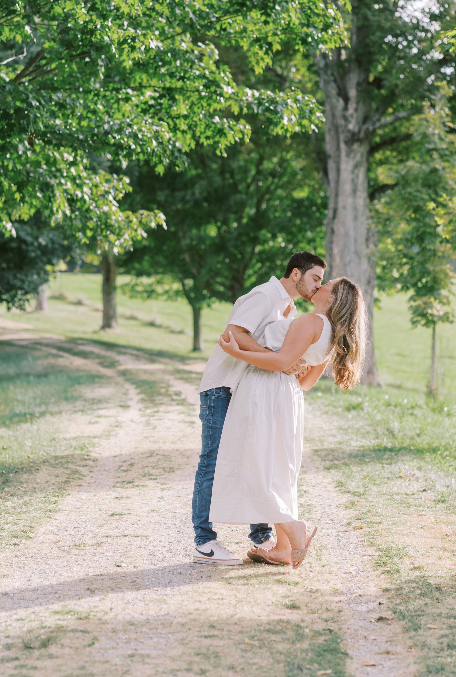 A couple shares playful moments along a tree-lined dirt path on a sunny day.