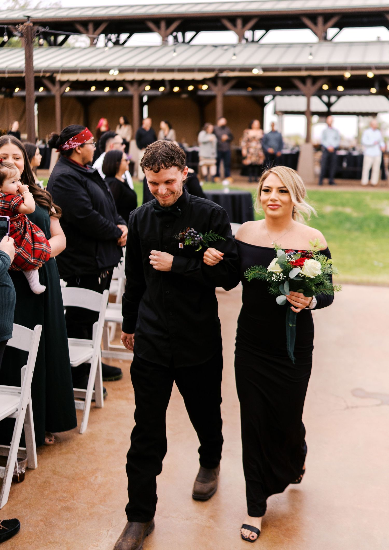 Members of a wedding party walk down the aisle at an indoor-outdoor venue decorated with chairs and hanging lights.