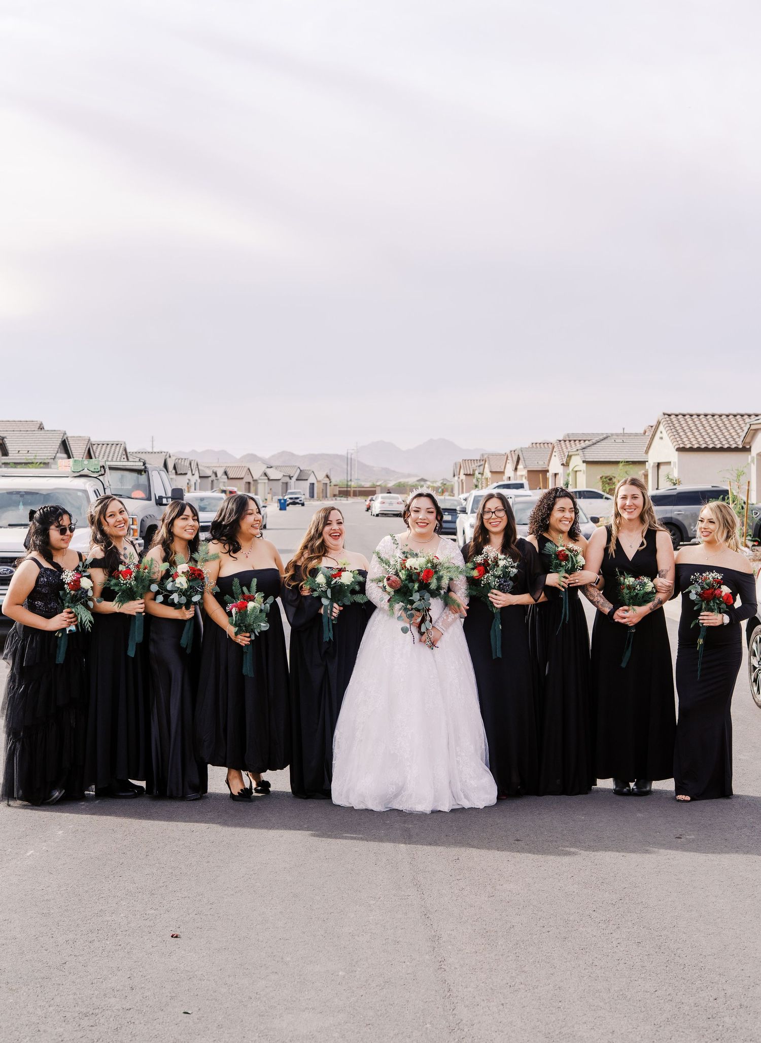 Wedding party in elegant black bridesmaid dresses pose with bride in white gown on a residential street at sunset.
