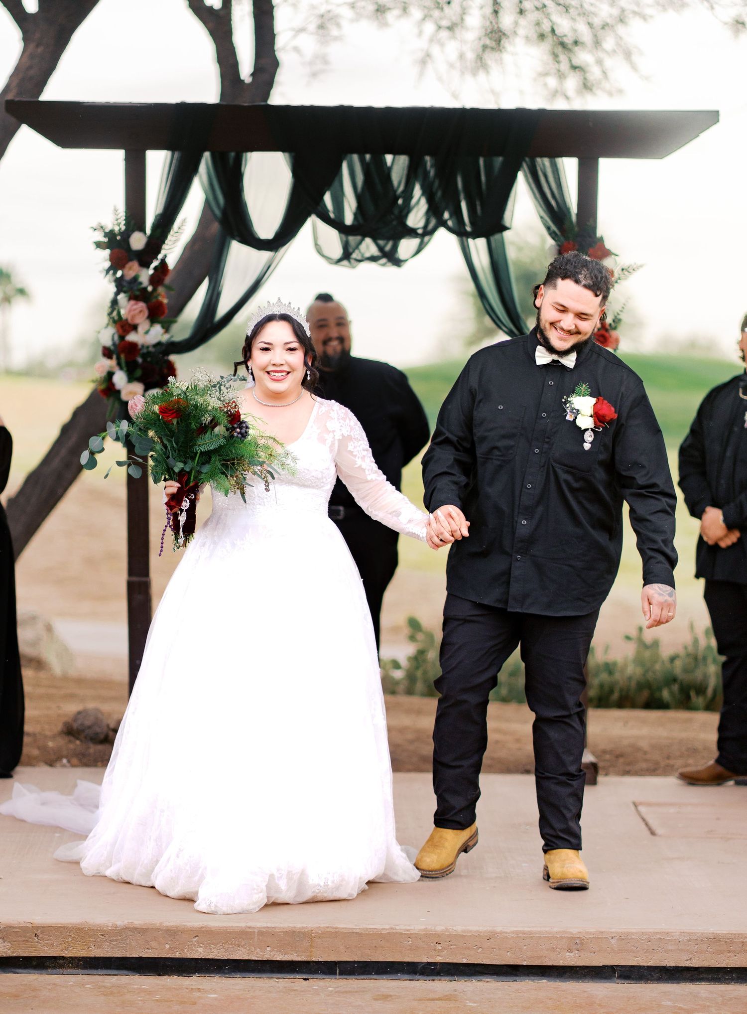 A bride and groom hold hands during their outdoor wedding ceremony under a rustic wooden arch with draped green fabric.