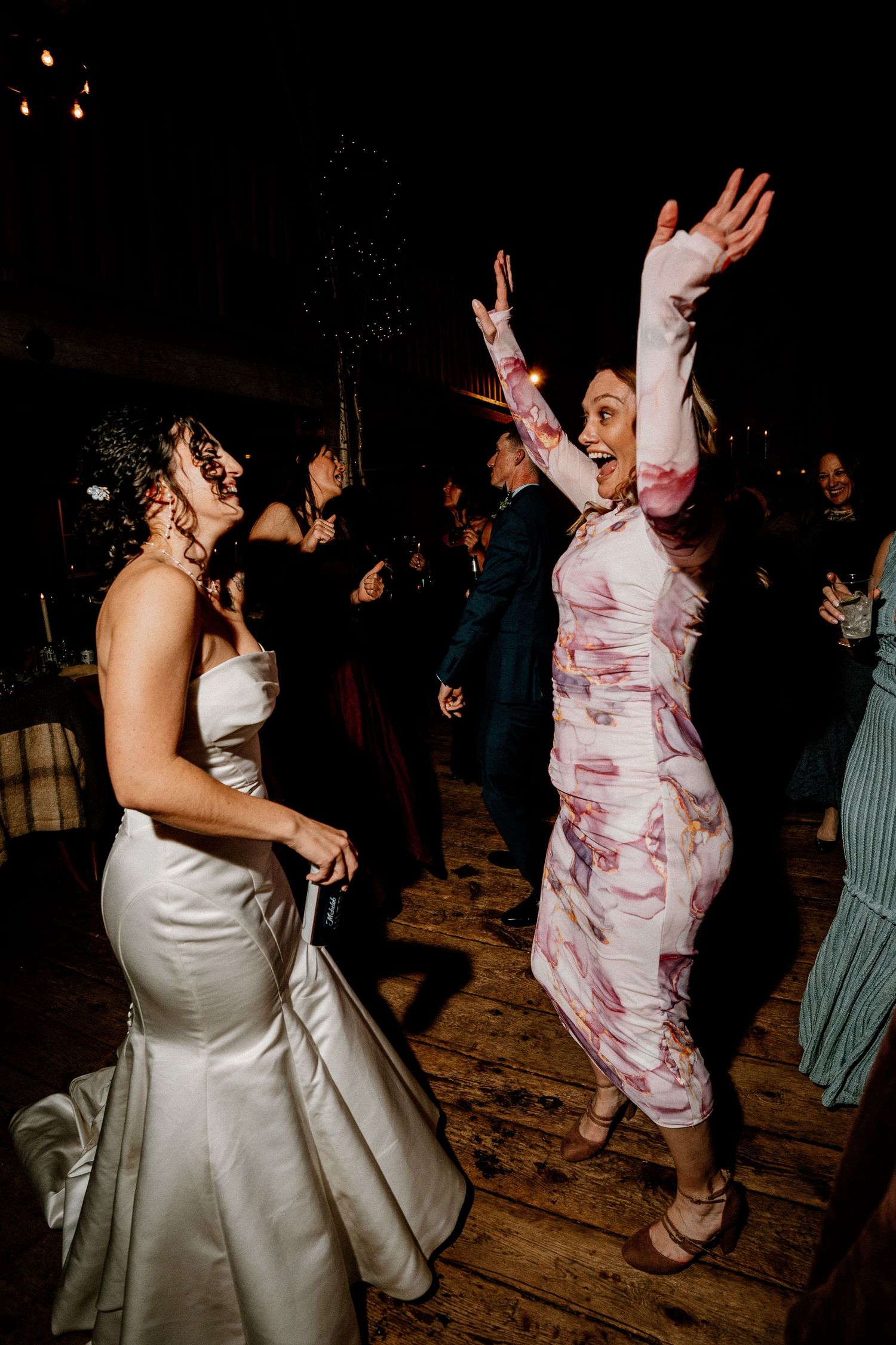 People dancing and celebrating at a wedding reception on a wooden floor, with one wearing white and another in pink floral.