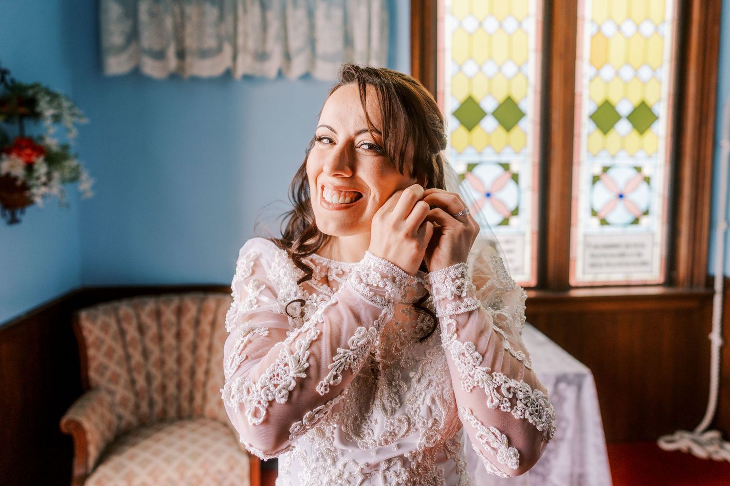 Bride in delicate lace dress laughs joyfully while getting ready in a room with stained glass windows.