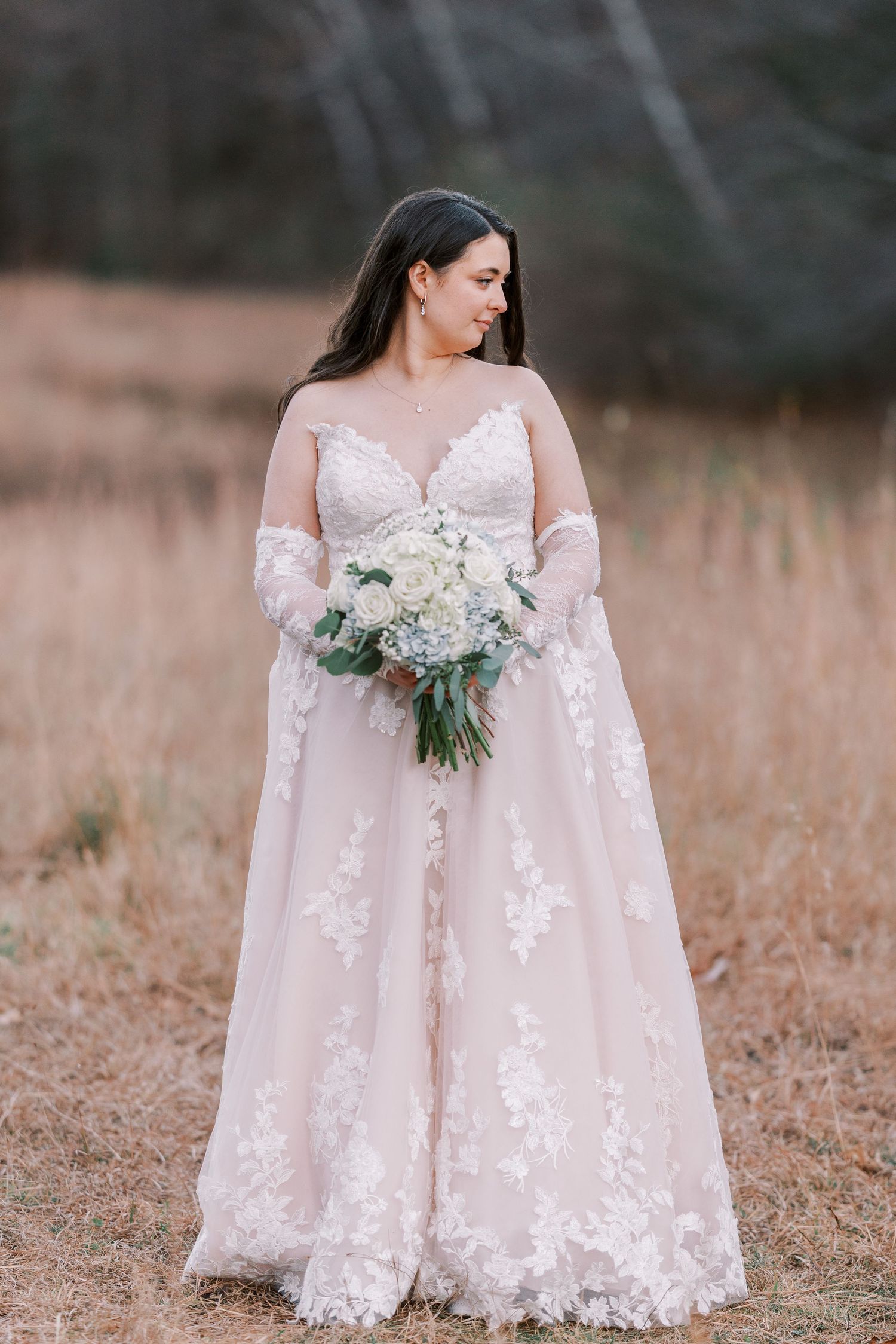 Bride in flowing lace wedding gown with sheer sleeves holds white floral bouquet while standing in autumn field.