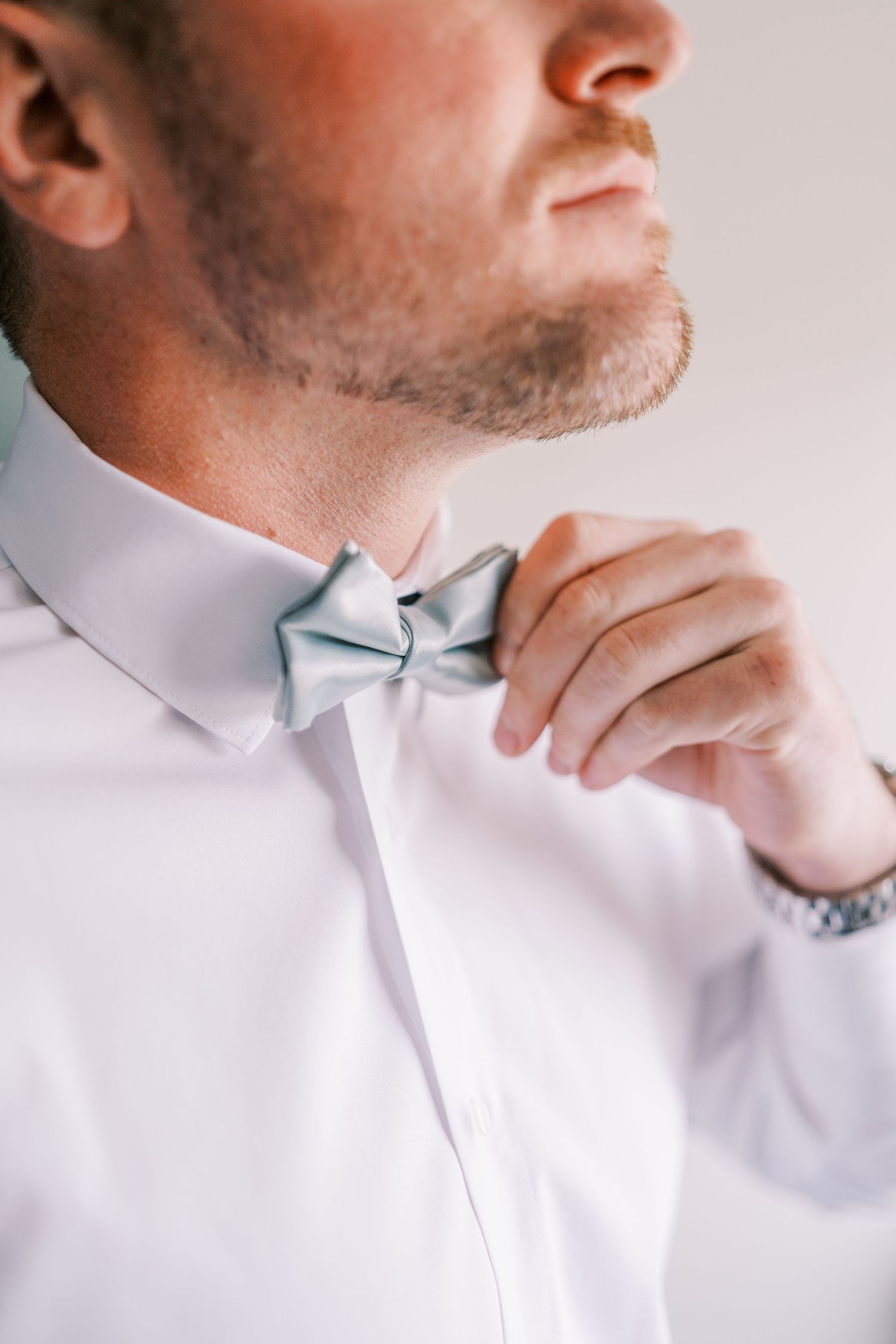 A person wearing a crisp white dress shirt adjusts their light blue bowtie while getting ready.