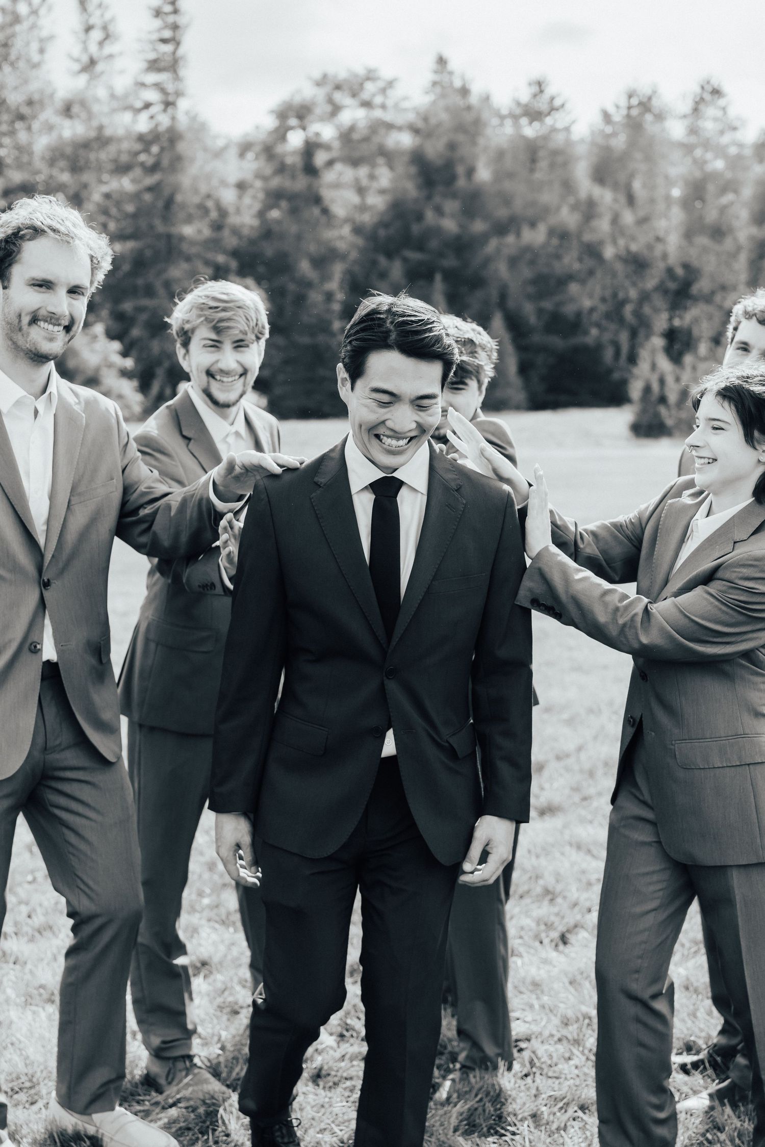 Groomsmen in dark suits laugh and embrace while standing in a line outdoors.