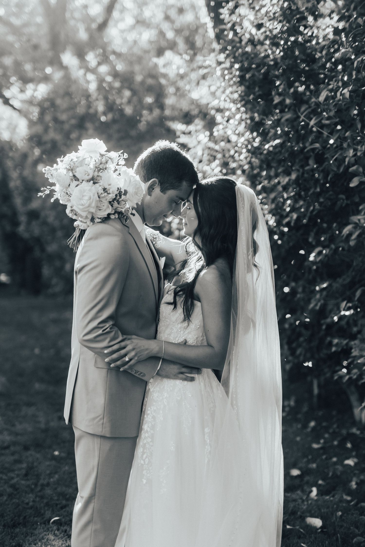 A romantic black and white wedding photo showing a couple embracing outdoors among garden greenery.