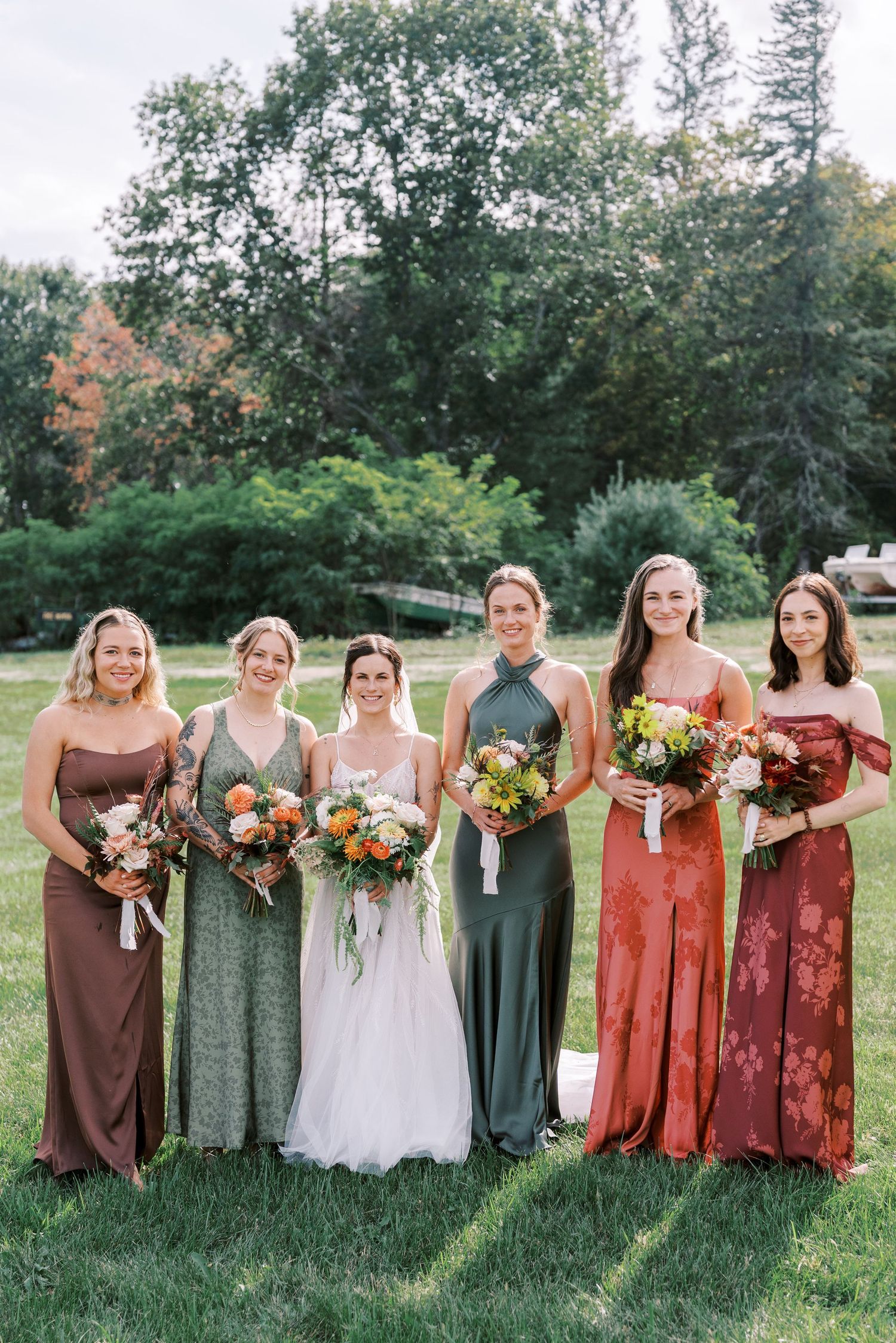 Bridesmaids line up in autumn-toned dresses holding colorful bouquets in outdoor setting.