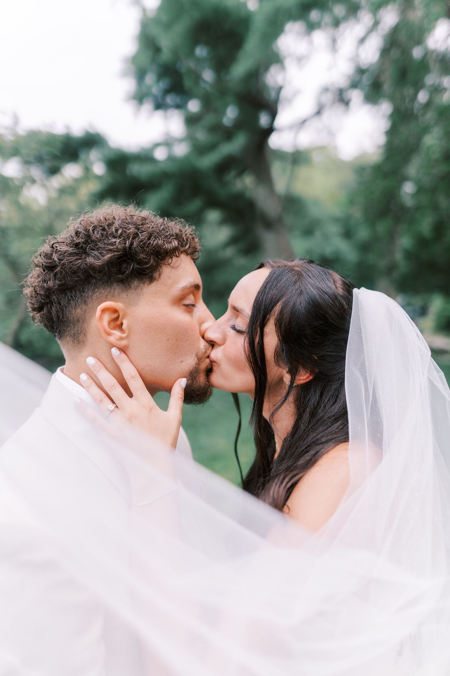 Romantic wedding portrait with flowing veil creating ethereal atmosphere in outdoor garden setting.