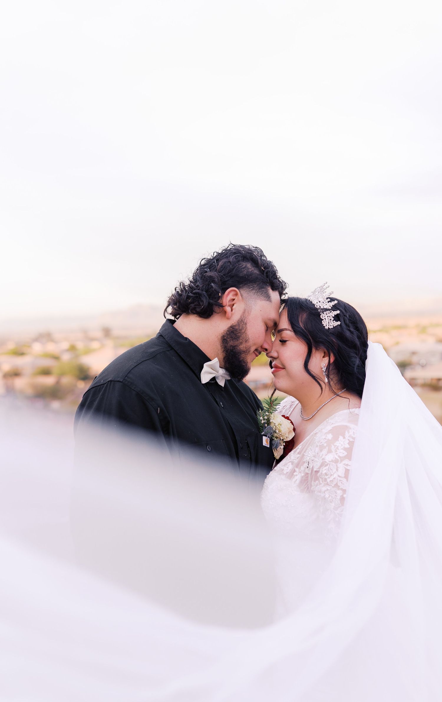 A romantic wedding couple shares an intimate moment against a desert landscape as the bride's veil flows in the wind.
