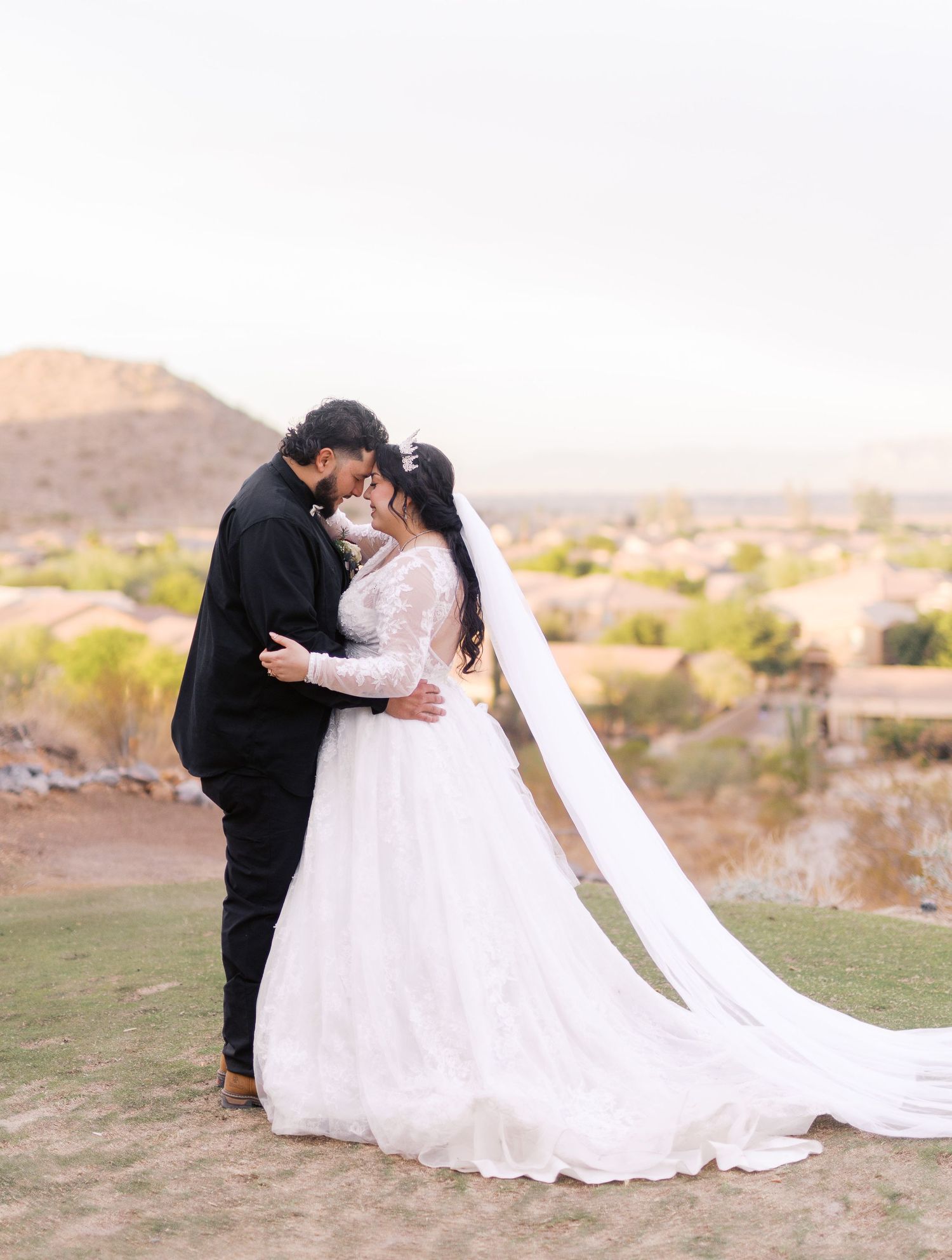 A couple in wedding attire share an intimate embrace on a hilltop overlooking desert landscape at sunset.