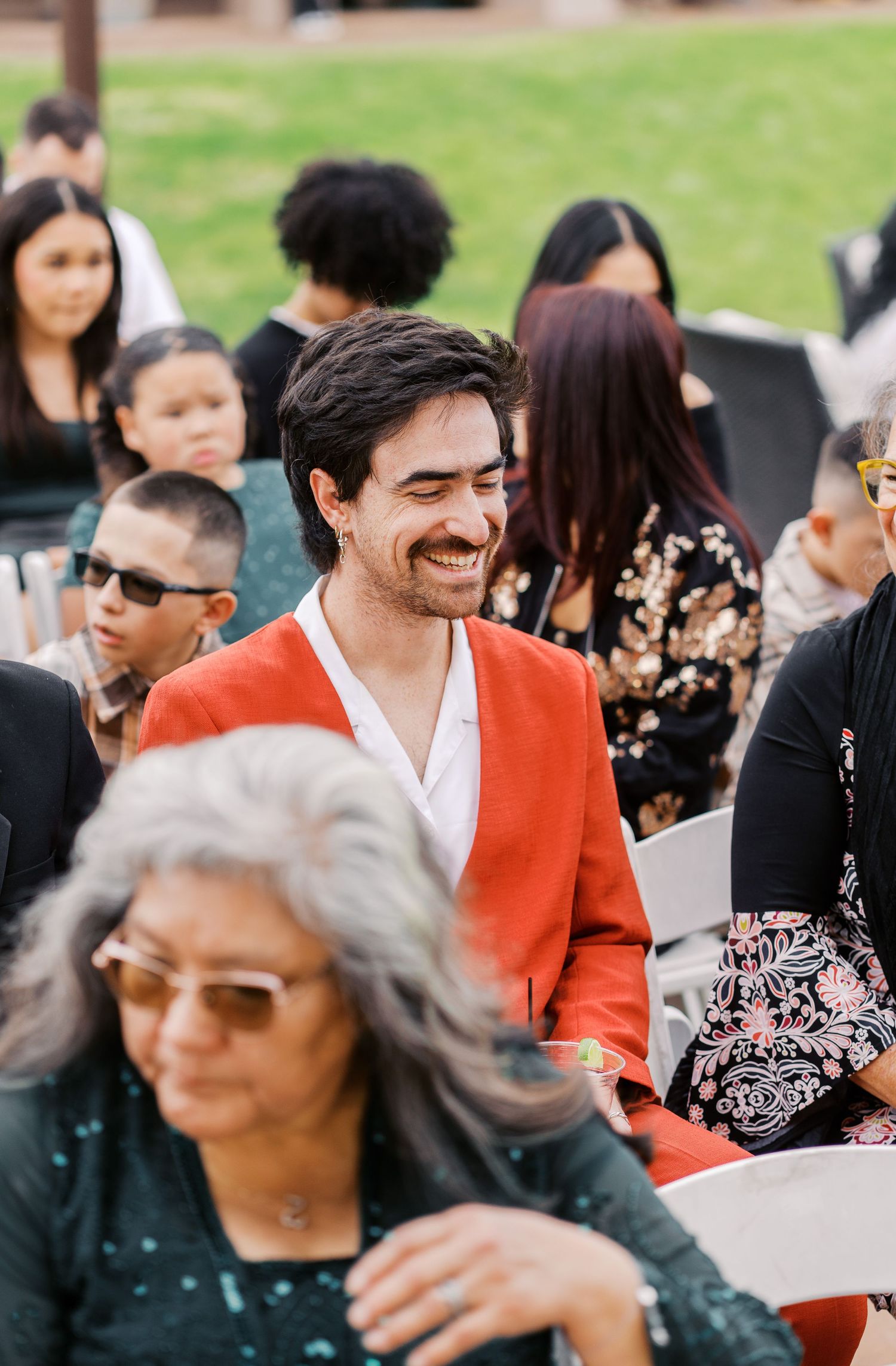 A crowd gathers outdoors with attendees in vibrant attire including a bright orange jacket at a social event.