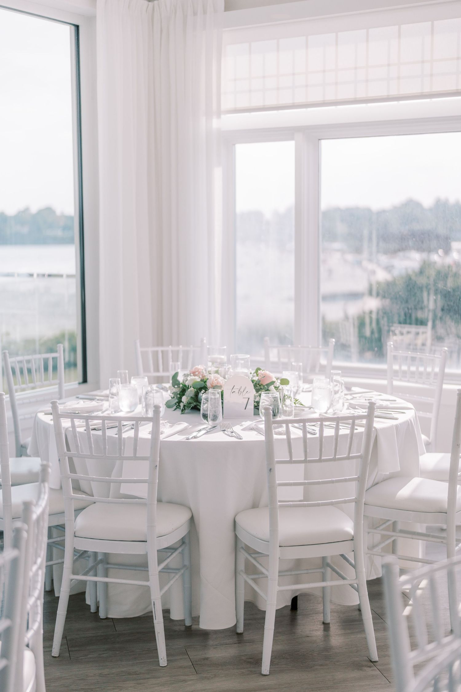 All-white wedding reception venue with elegant table settings and floor-to-ceiling windows overlooking water views.