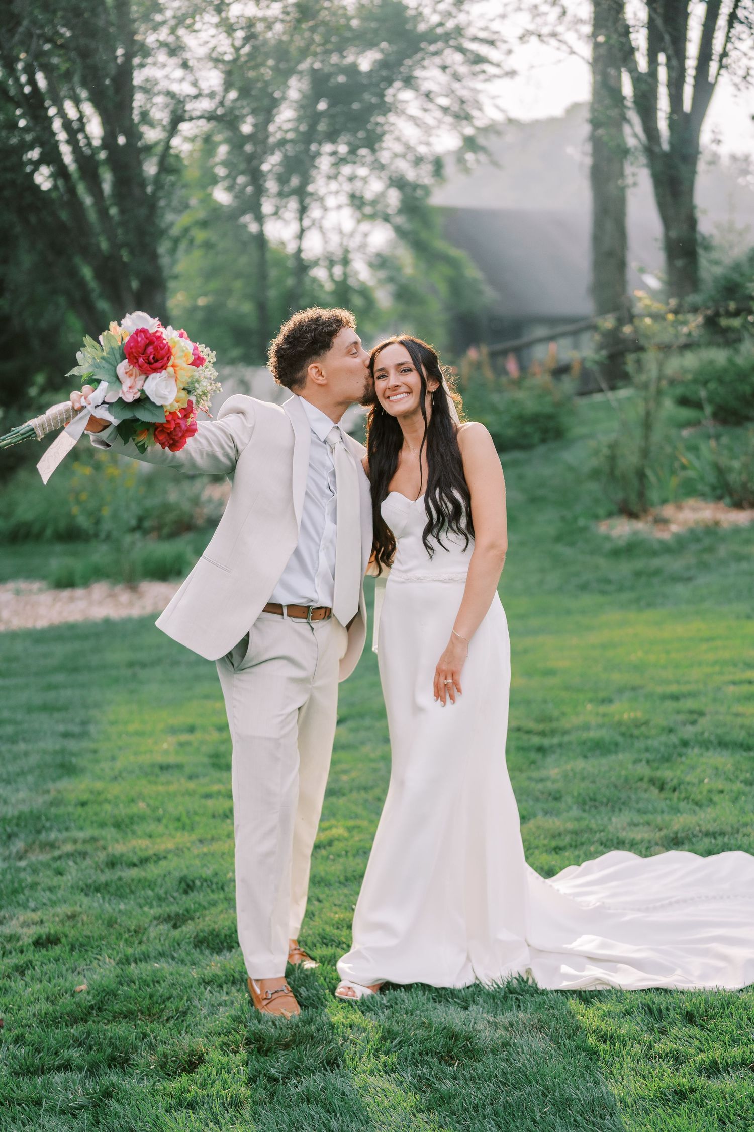 A couple poses playfully on their wedding day with a bright floral bouquet against green scenery.