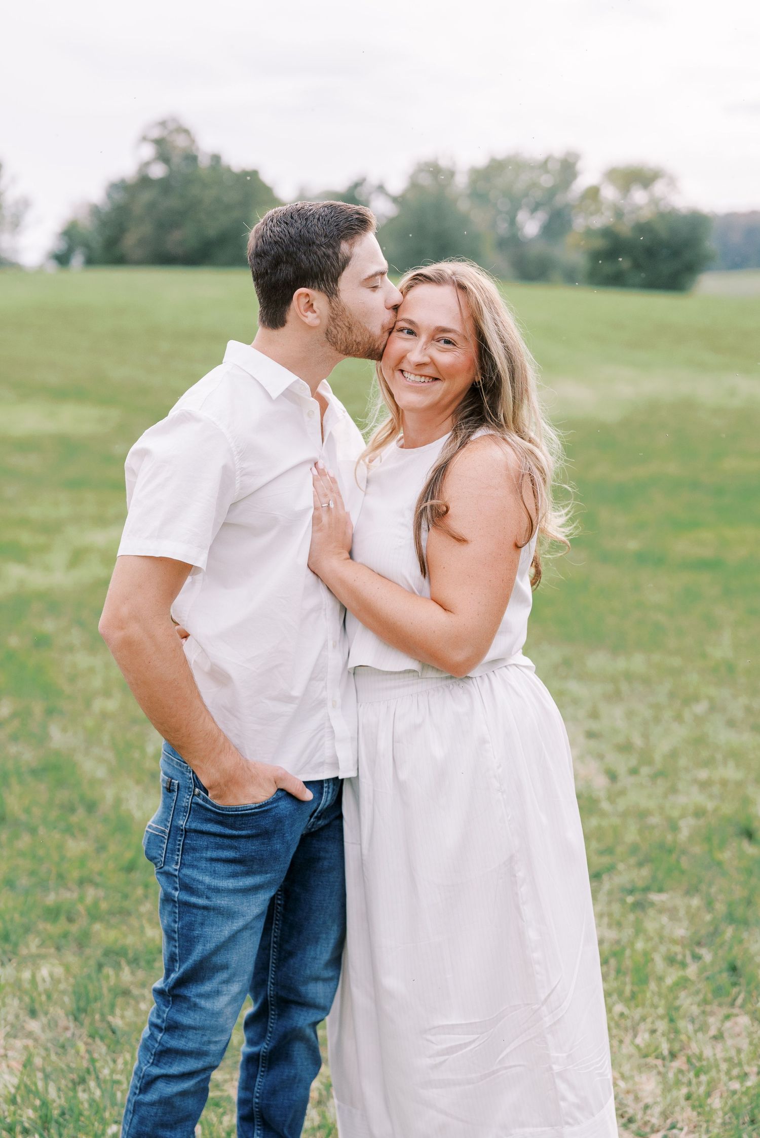 Affectionate couple in white outfits sharing tender moment in sunny meadow.