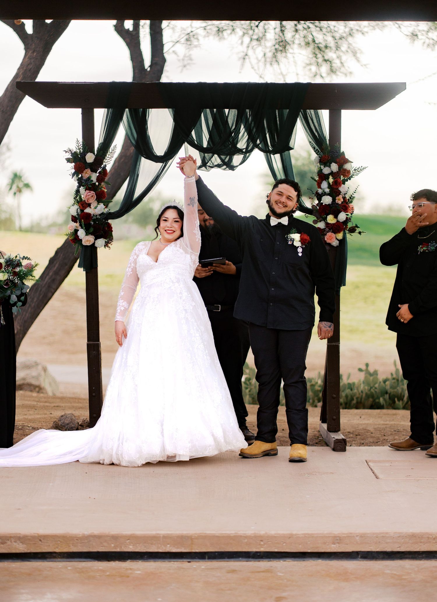 A joyful wedding ceremony taking place under a wooden arch decorated with dark green drapes and pink floral arrangements.
