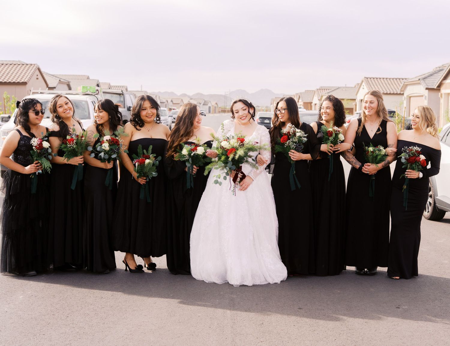 Wedding party in black bridesmaid dresses and white bridal gown pose together on a sunny residential street.