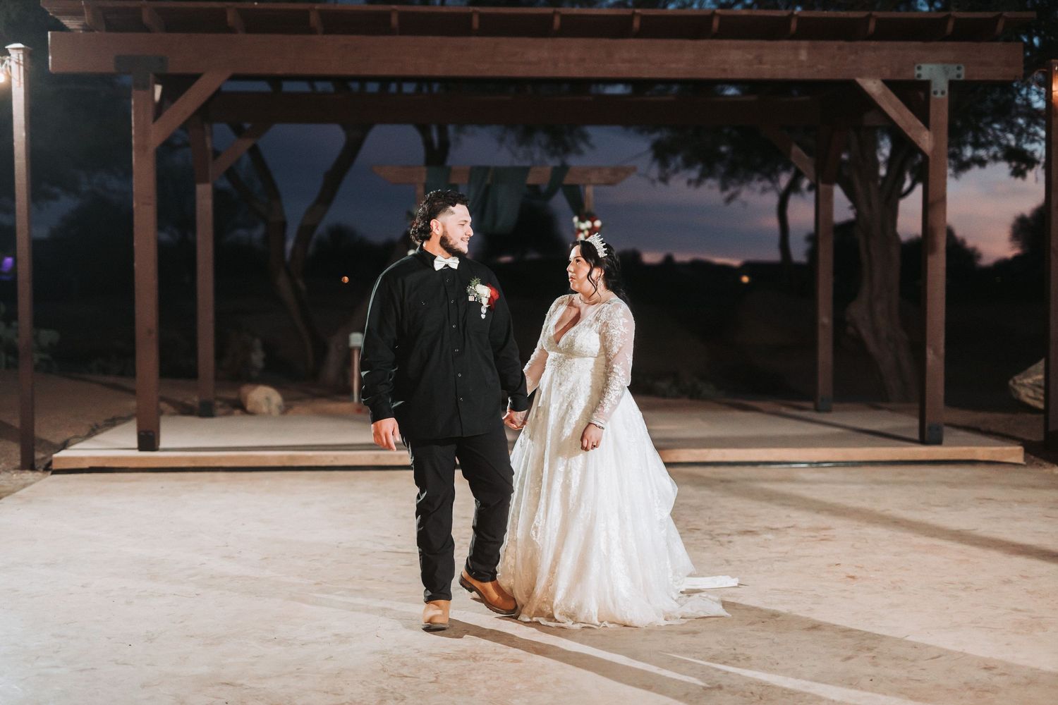 A couple in wedding attire stands beneath a wooden pergola at dusk during their outdoor celebration.